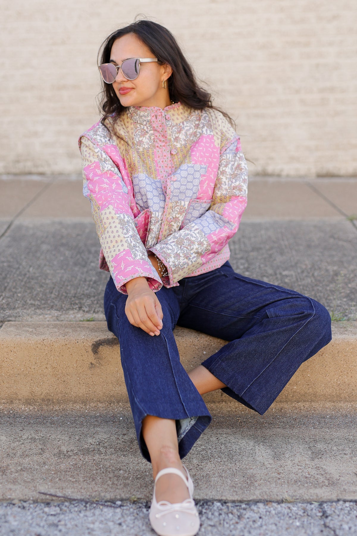 A woman wearing a colorful patchwork quilted jacket with pink, blue, and gold patterns, paired with dark blue cropped jeans and white flats, sitting on concrete steps against a light brick wall.