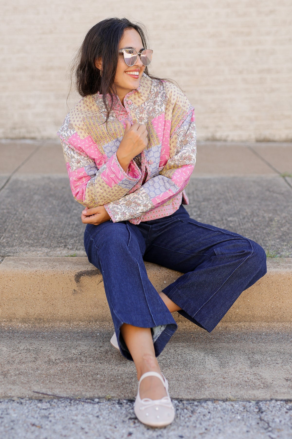 A woman wearing a colorful patchwork quilted jacket, dark blue cropped jeans, and white flats sits on concrete steps against a beige brick wall.