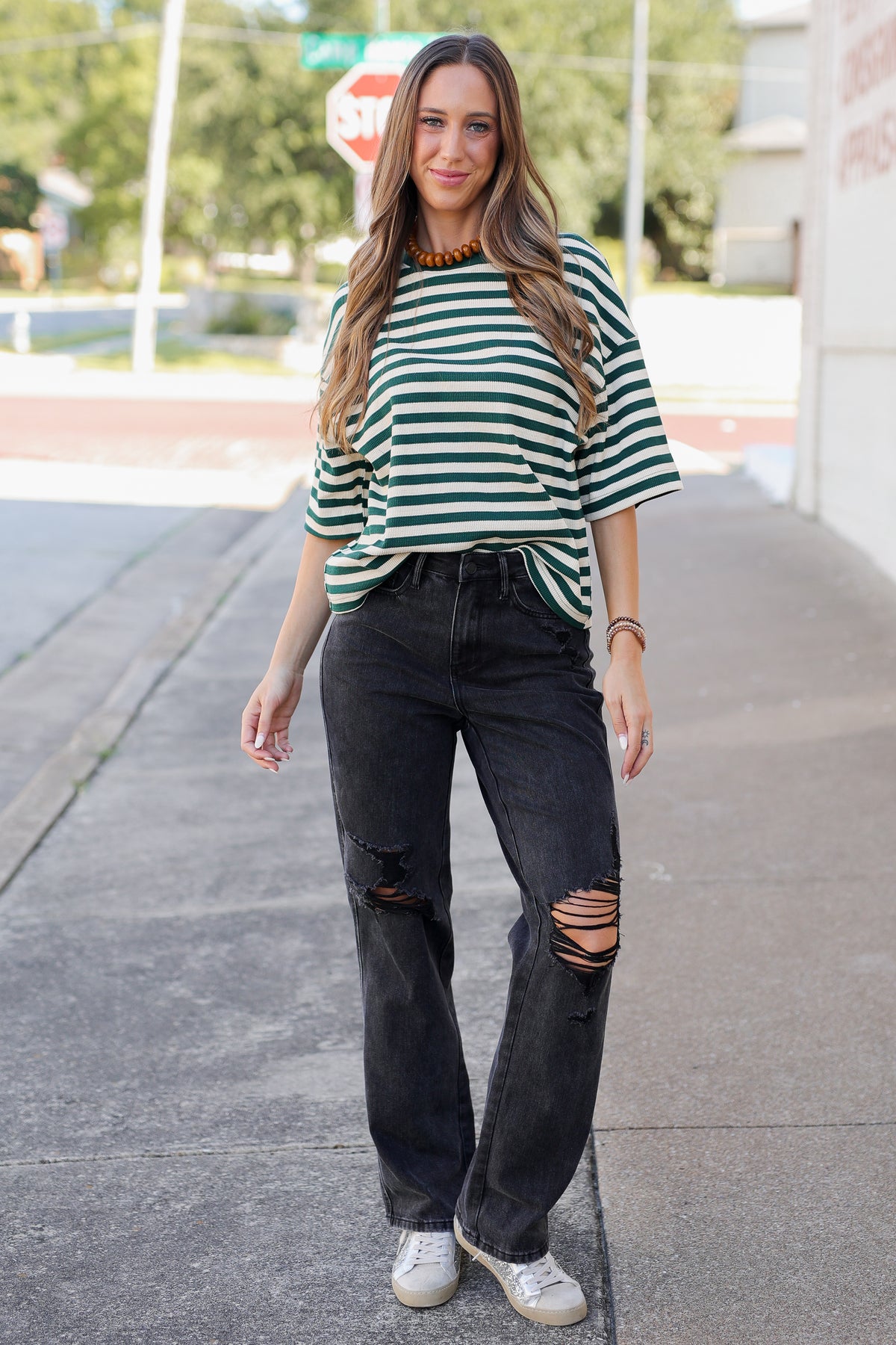A woman stands on a sidewalk wearing a green and white striped short-sleeve top, black distressed jeans, and white sneakers.