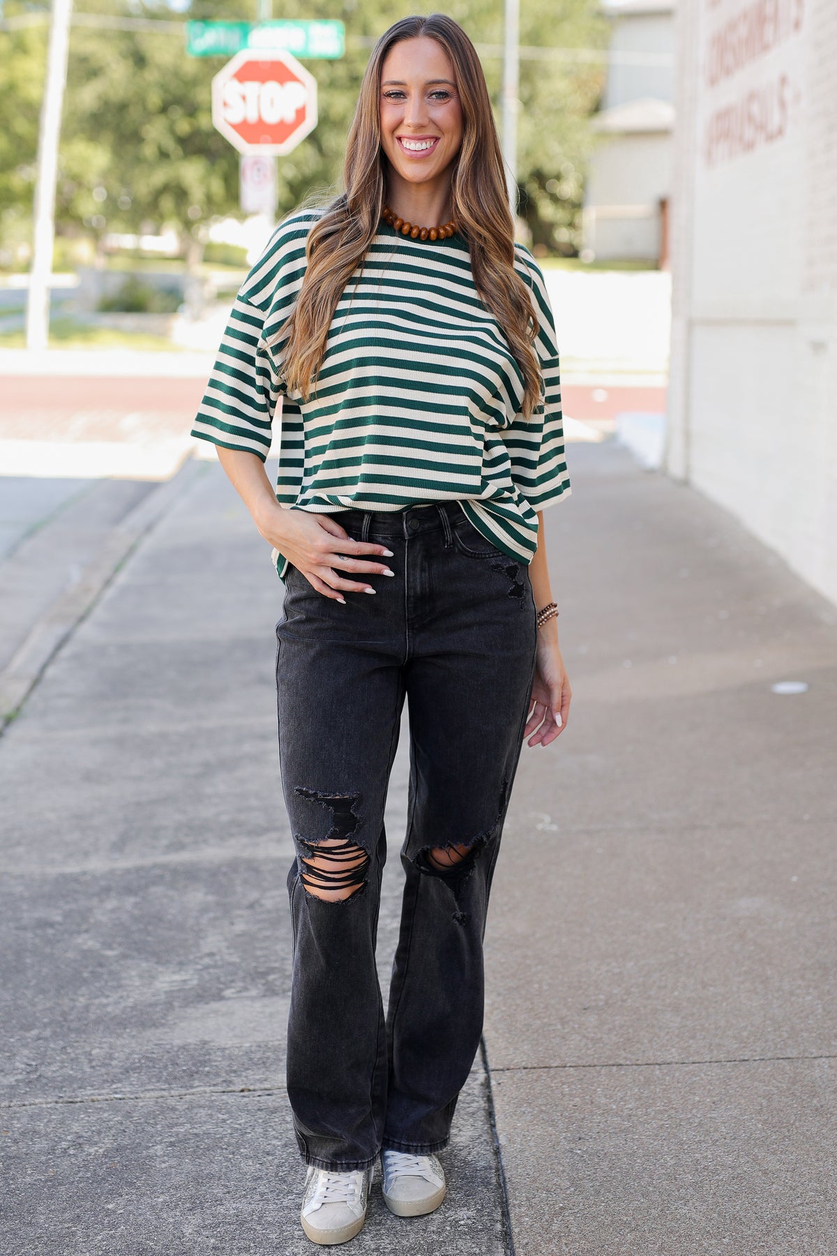 A woman stands on a sidewalk wearing a green and white striped short-sleeve top, black ripped jeans, and white sneakers.