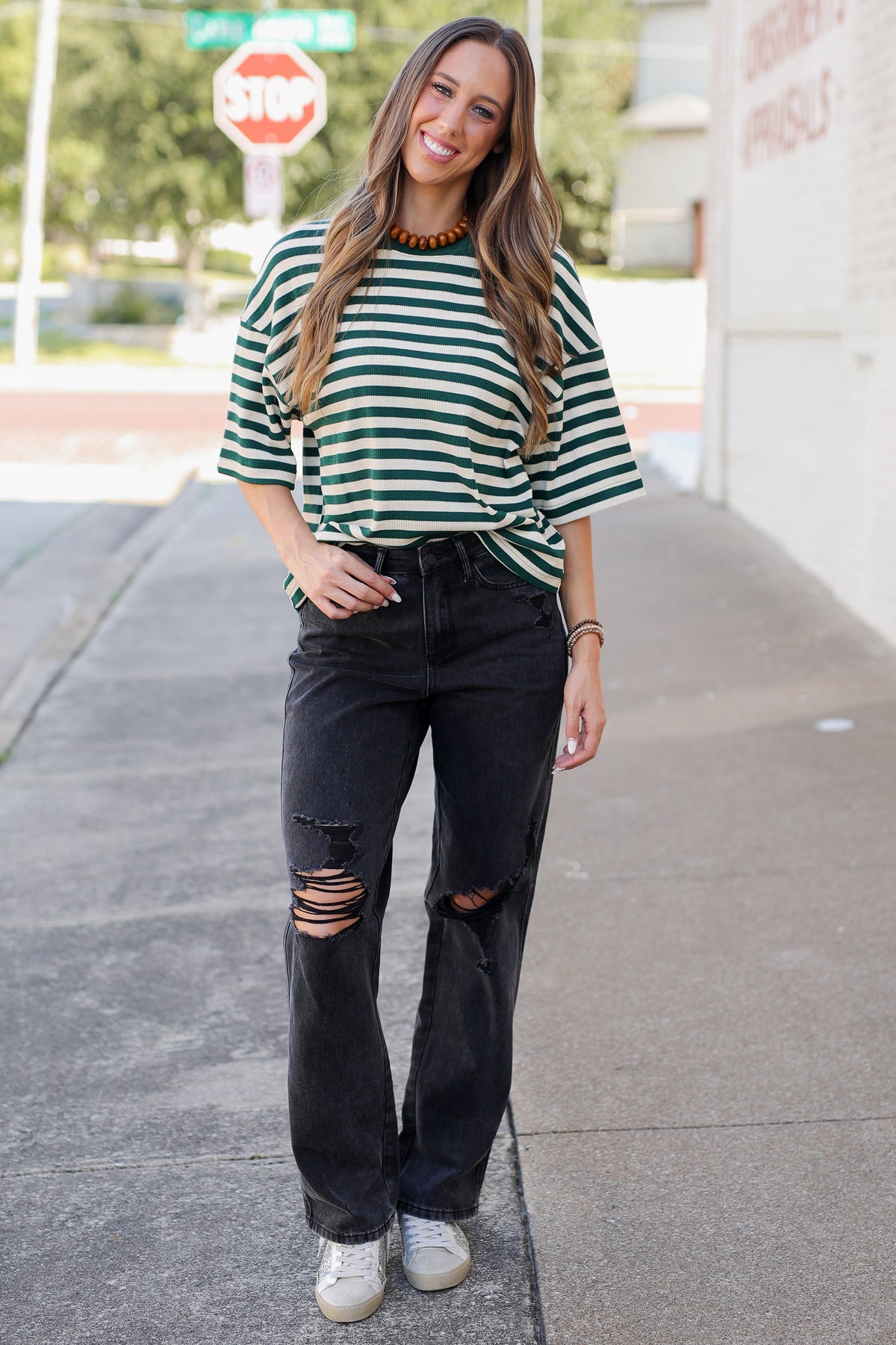 A woman stands on a sidewalk wearing an oversized green and white striped t-shirt, black ripped jeans, and white sneakers.