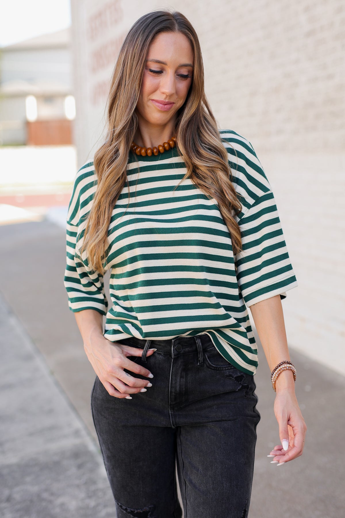 A woman wearing a green and white striped short-sleeve top with black jeans, standing outdoors against a light-colored brick wall.