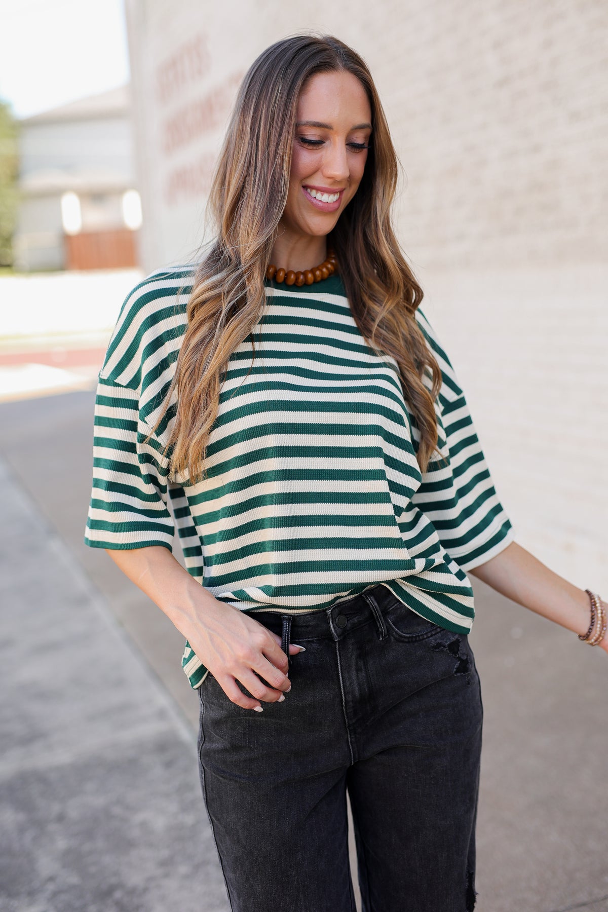 A woman wearing a green and white striped short-sleeve t-shirt with dark gray jeans, standing outdoors against a light-colored brick wall.