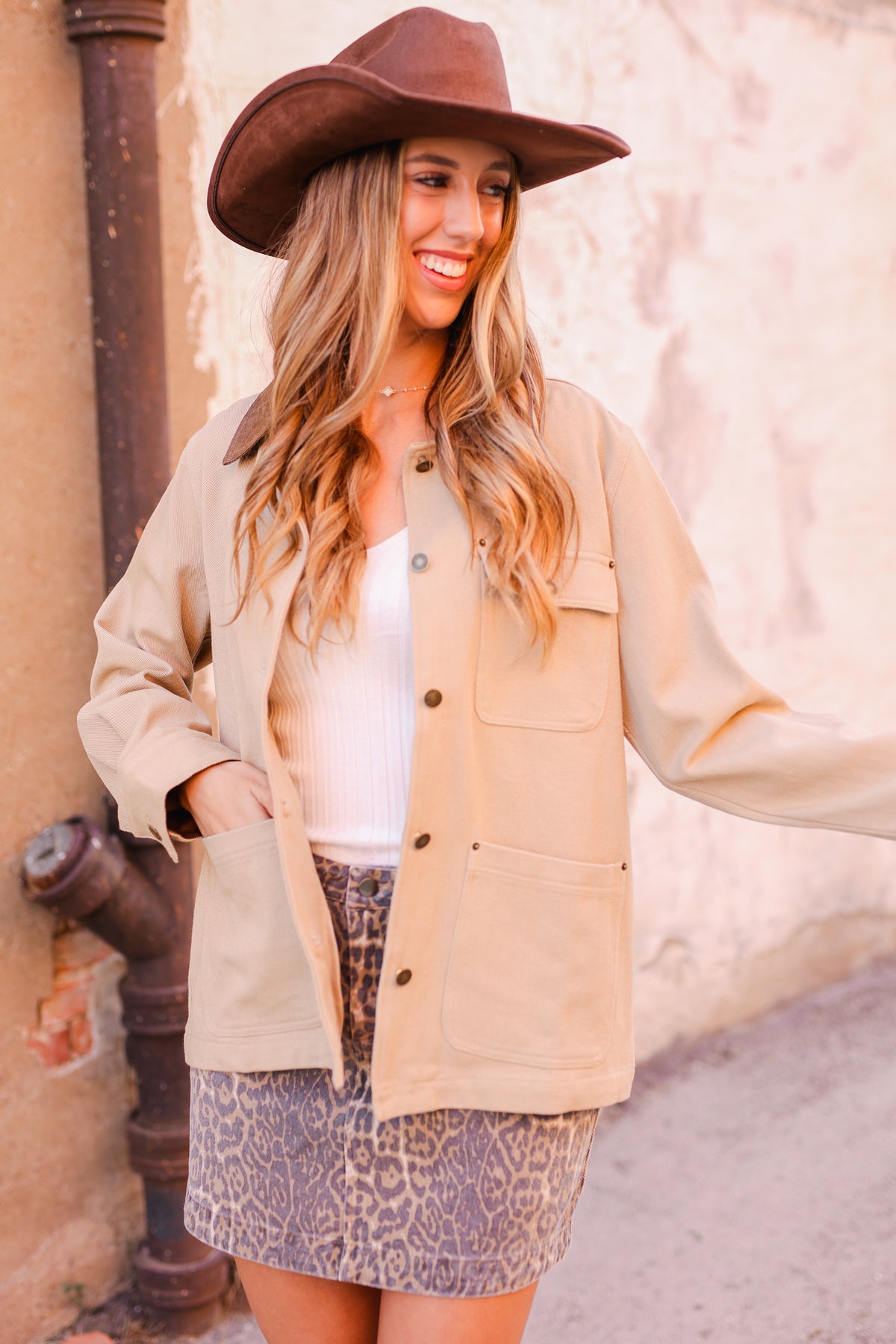 Woman wearing a beige coat, white top, and patterned skirt with a brown hat against a beige wall.