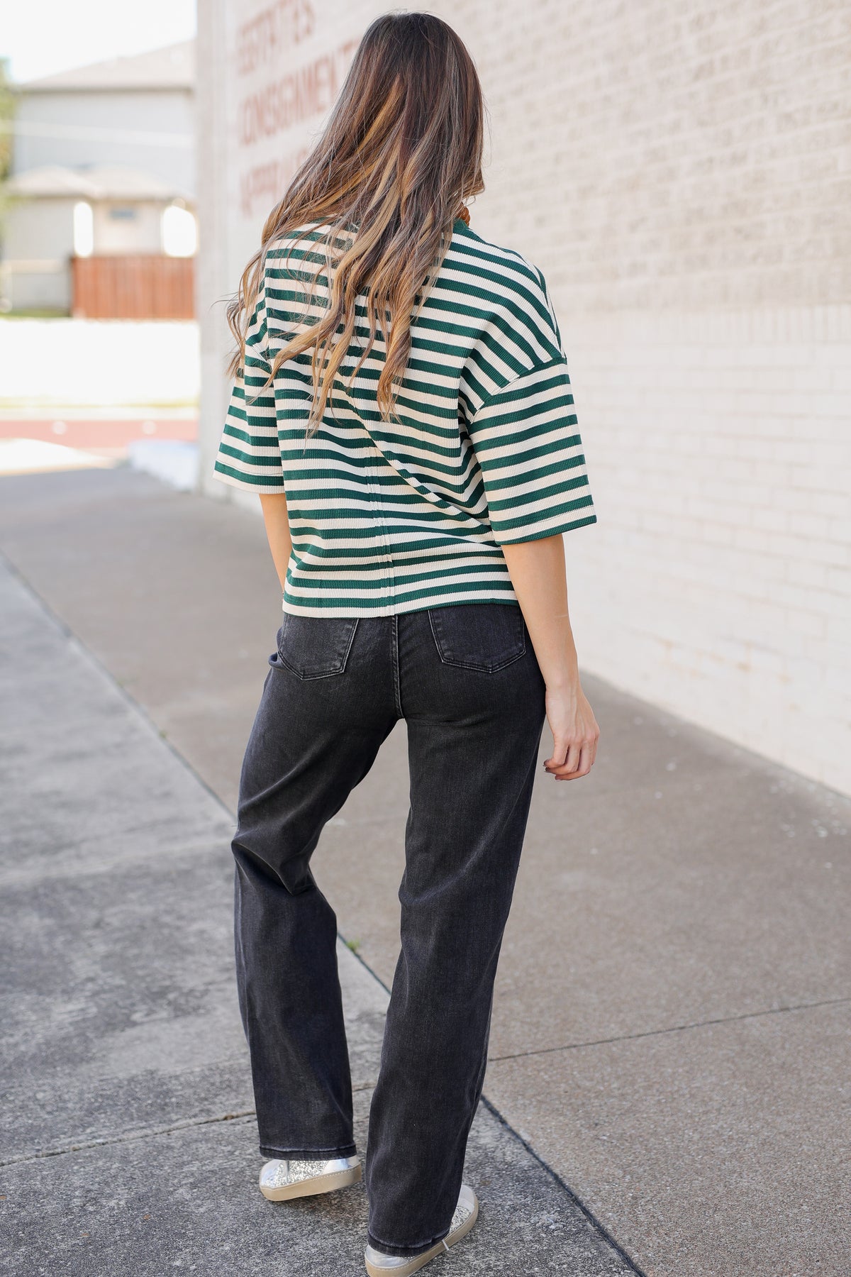 A woman viewed from behind wearing a green and white striped short-sleeve top, dark charcoal jeans, and white sneakers, standing on a sidewalk next to a light brick wall.