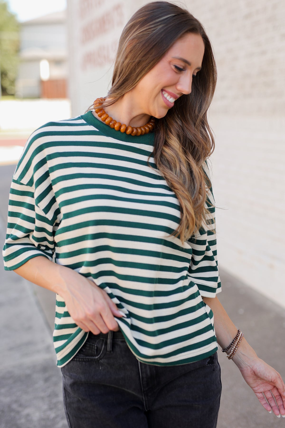 A woman wearing a green and white striped long-sleeve top with dark jeans, standing outdoors against a light brick wall.
