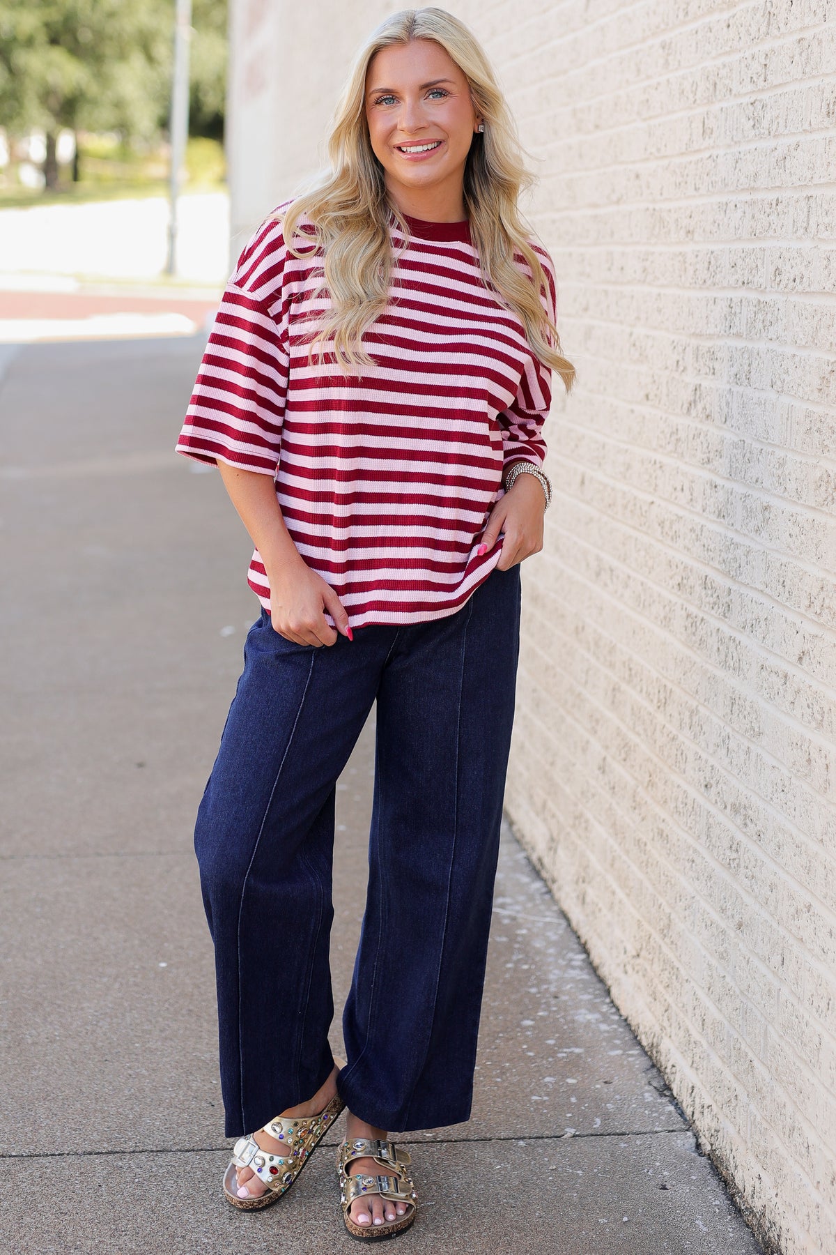 A woman stands outdoors against a light brick wall wearing a red and white horizontally striped short-sleeve top, dark navy wide-leg pants, and patterned sandals.