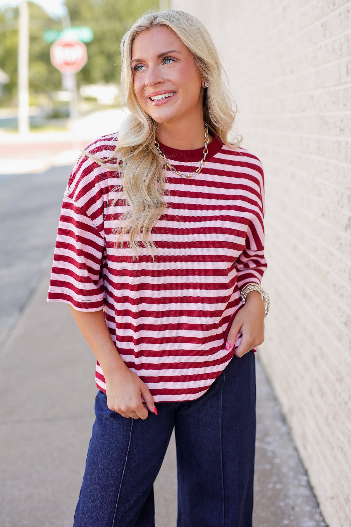 A woman wearing a red and white striped short-sleeve top and dark blue jeans, standing outdoors next to a light-colored brick wall.