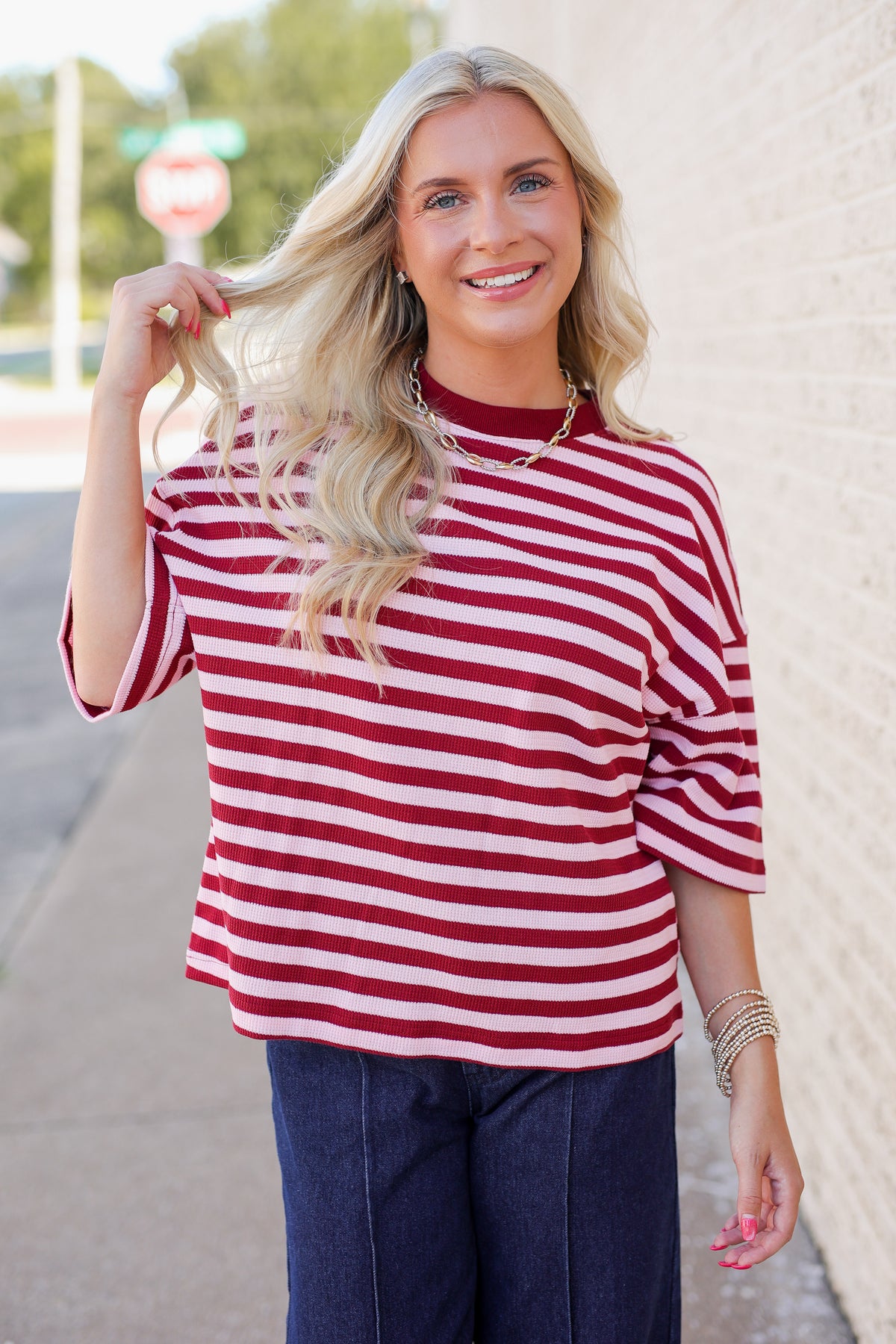 A smiling blonde woman wearing a red and white horizontally striped short-sleeve top with dark blue jeans, standing outdoors against a light brick wall.