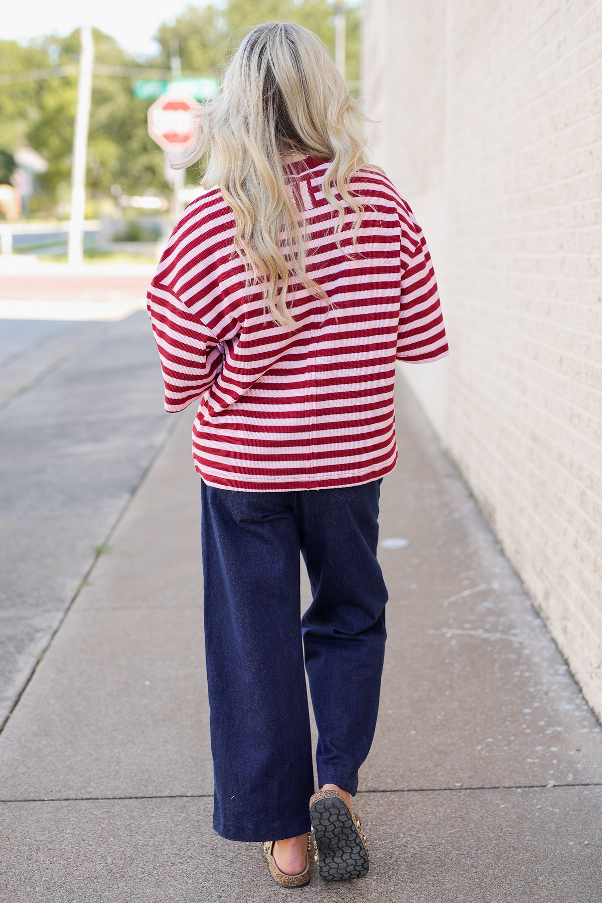 A woman viewed from behind walking on a sidewalk, wearing an oversized red and white striped shirt and dark navy wide-leg pants.