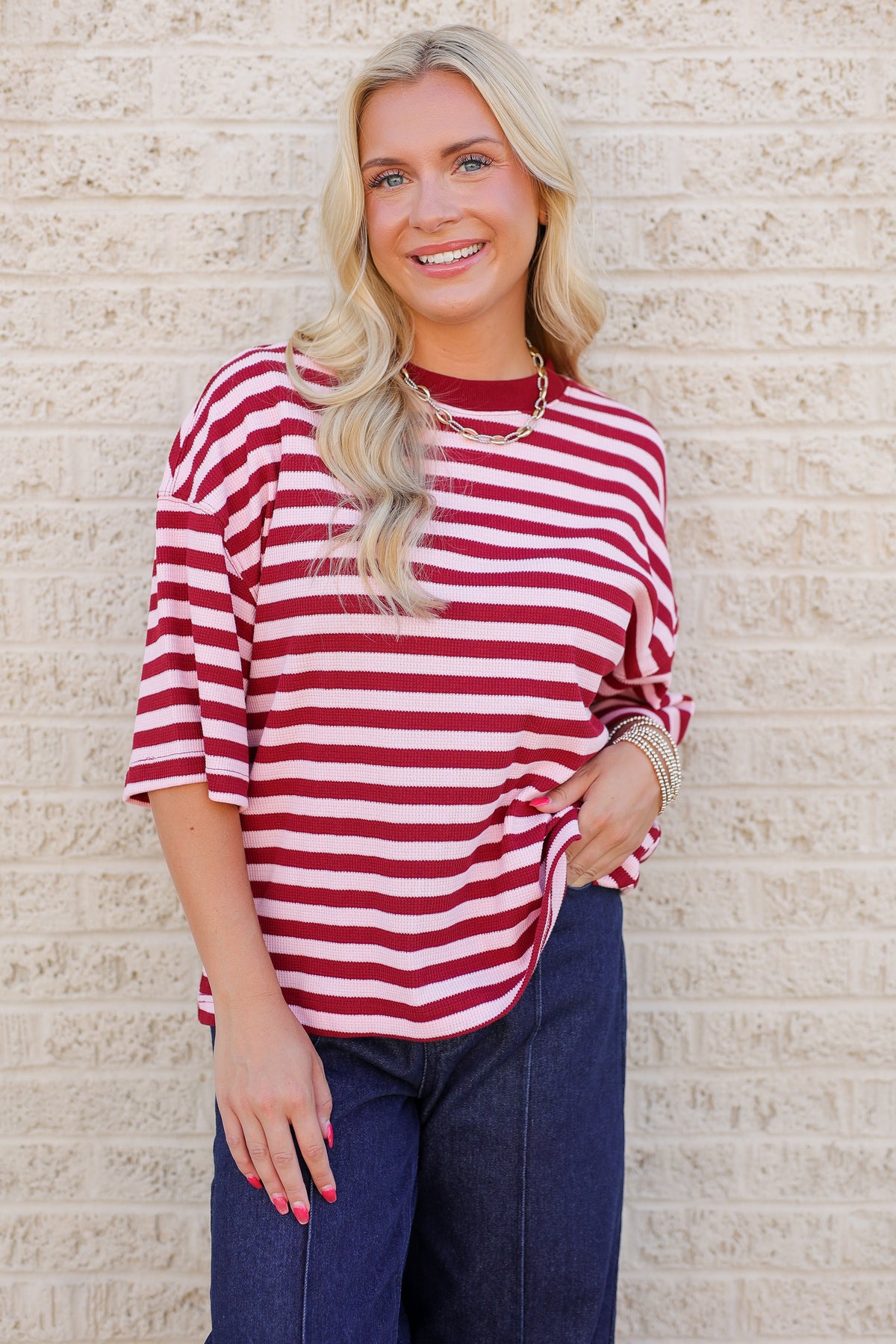 A woman with blonde hair wearing a red and white horizontally striped short-sleeve top and dark blue jeans, standing against a light beige brick wall.