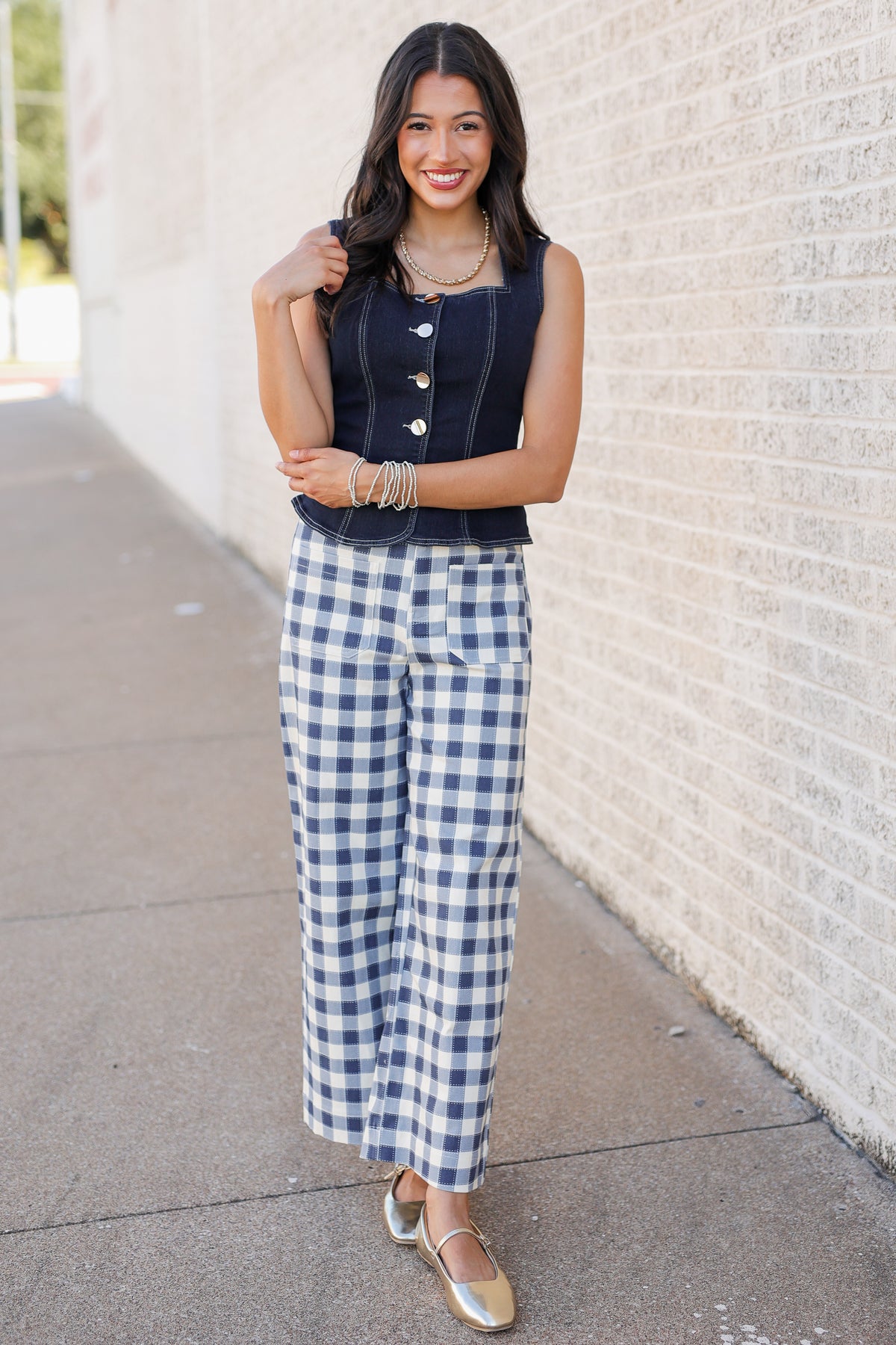 A woman stands outdoors against a light brick wall wearing a navy sleeveless top with gold buttons and blue-and-white gingham wide-leg pants, paired with metallic gold flats.