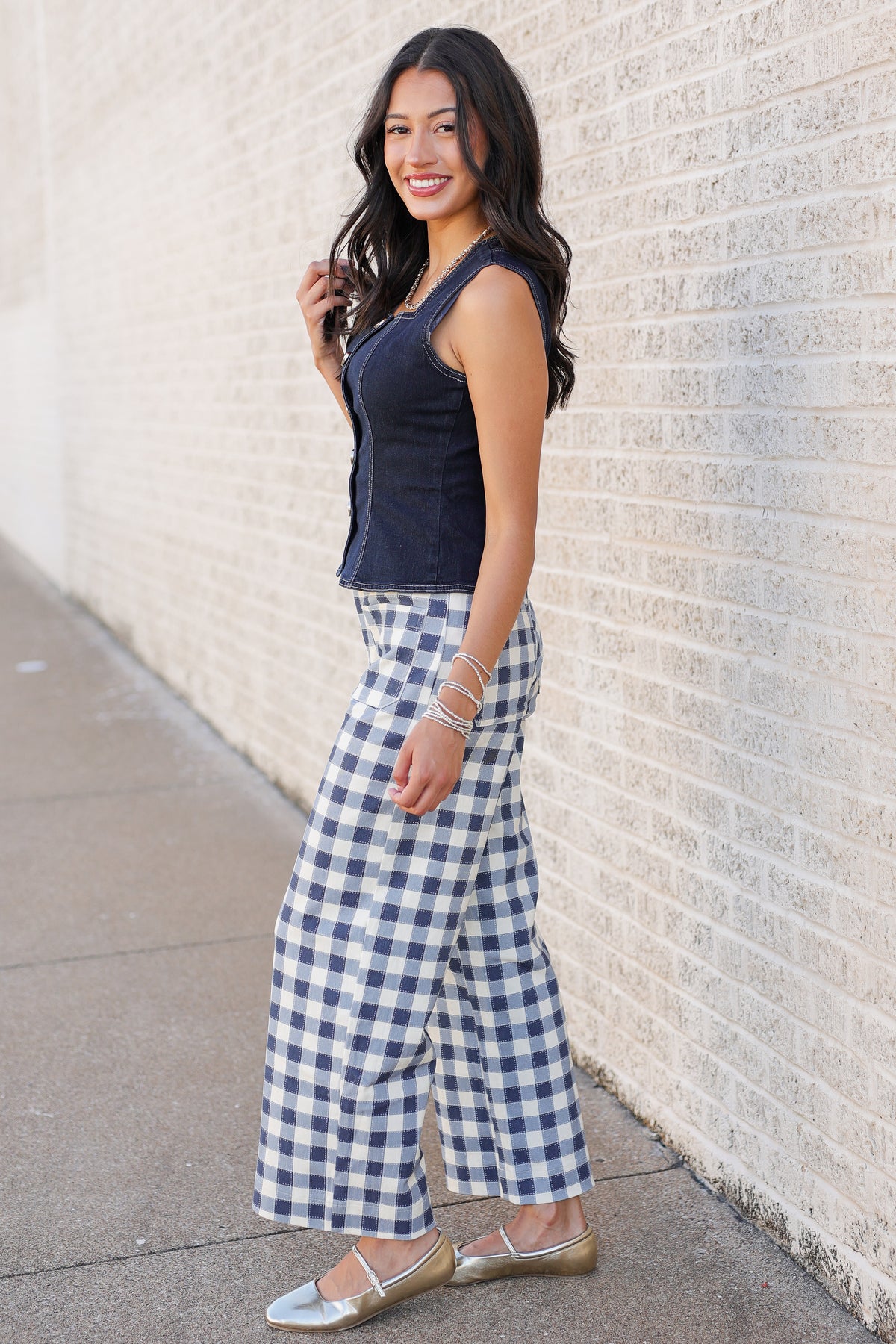 A woman posing outdoors against a light brick wall, wearing a navy blue sleeveless top and blue-and-white gingham wide-leg pants, paired with metallic gold flats.