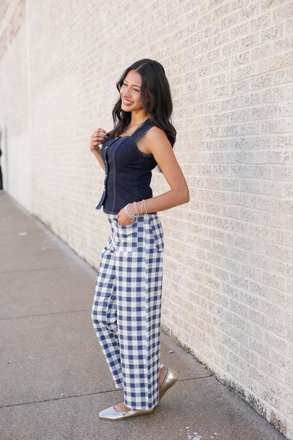 A woman posing outdoors against a light brick wall, wearing a navy sleeveless top and blue-and-white gingham wide-leg pants.