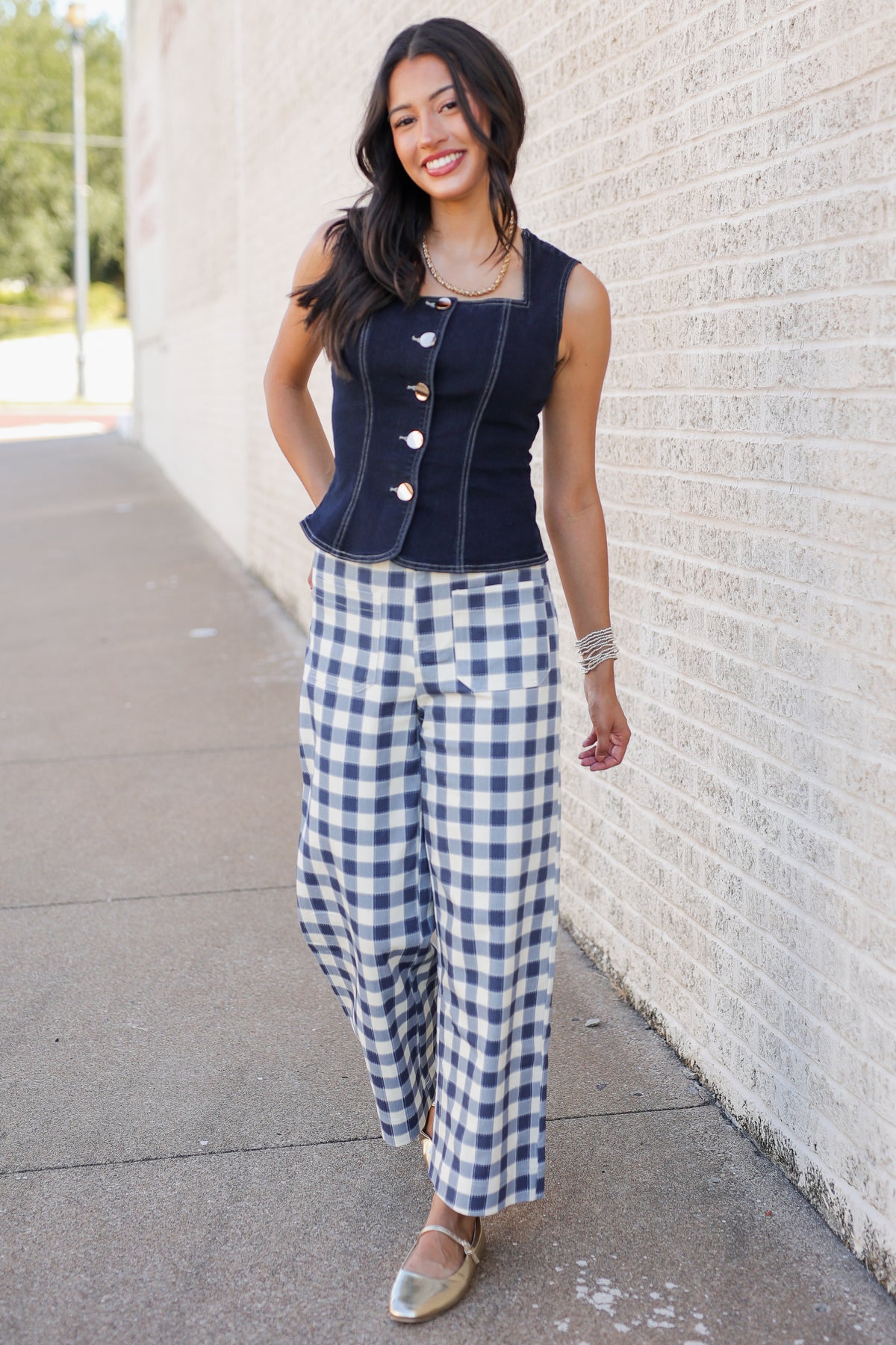 A woman stands outdoors against a light brick wall wearing a sleeveless navy top with gold buttons and blue-and-white gingham cropped pants.