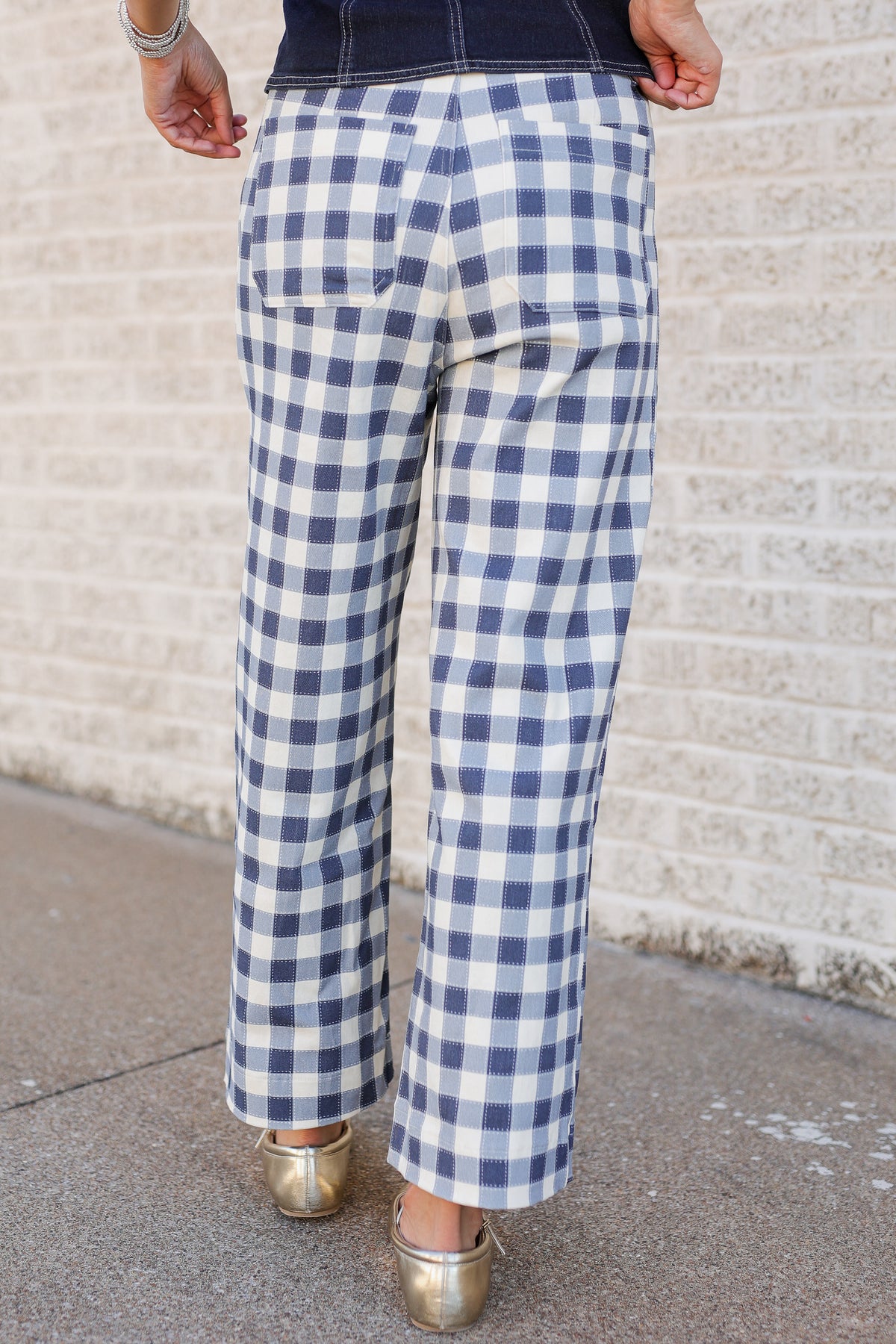 A person shown from the back wearing blue and white gingham cropped pants, a dark navy top, and metallic gold heels, standing against a light brick wall.