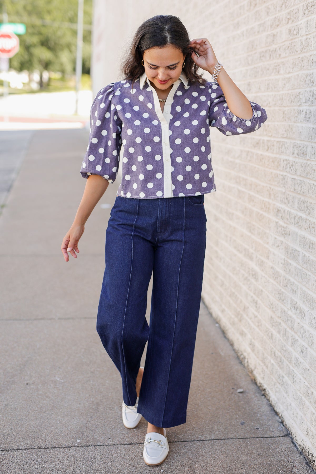 A woman walking on a sidewalk next to a light brick wall, wearing a lavender and white polka dot cropped blouse with navy blue wide-leg pants and white slip-on shoes.
