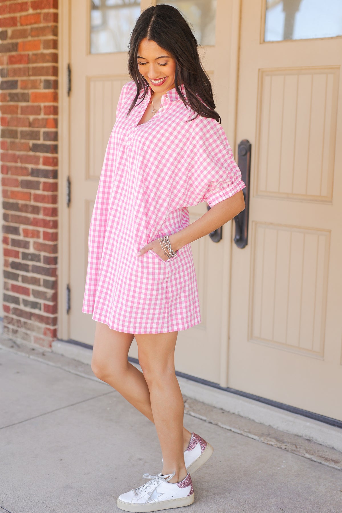 Woman wearing a pink checkered dress and white sneakers standing in front of a door.