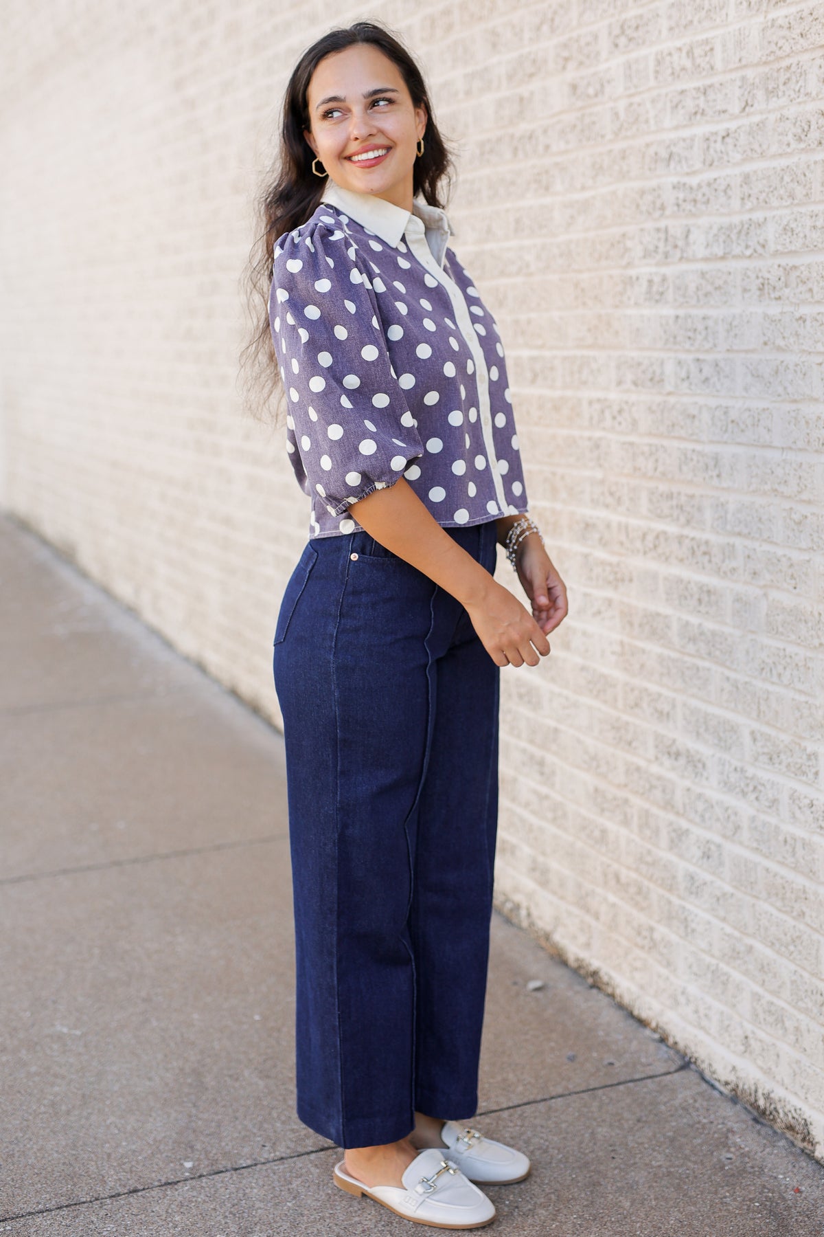 A woman stands outdoors against a light brick wall wearing a lavender blouse with white polka dots and a white collar, navy wide-leg cropped pants, and white loafers.