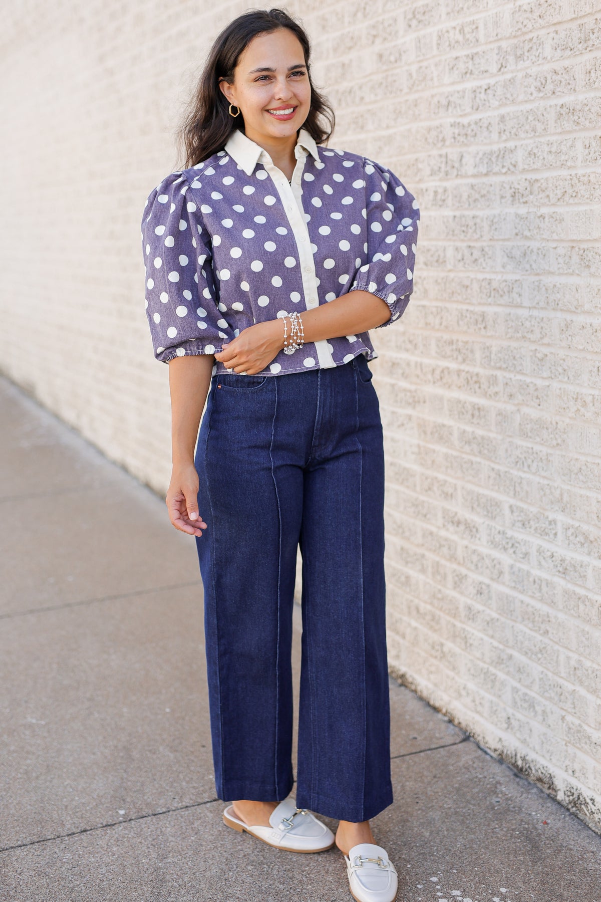 A woman stands outdoors against a light brick wall wearing a purple and white polka dot blouse with puffed sleeves, dark blue wide-leg pants, and white slip-on shoes.