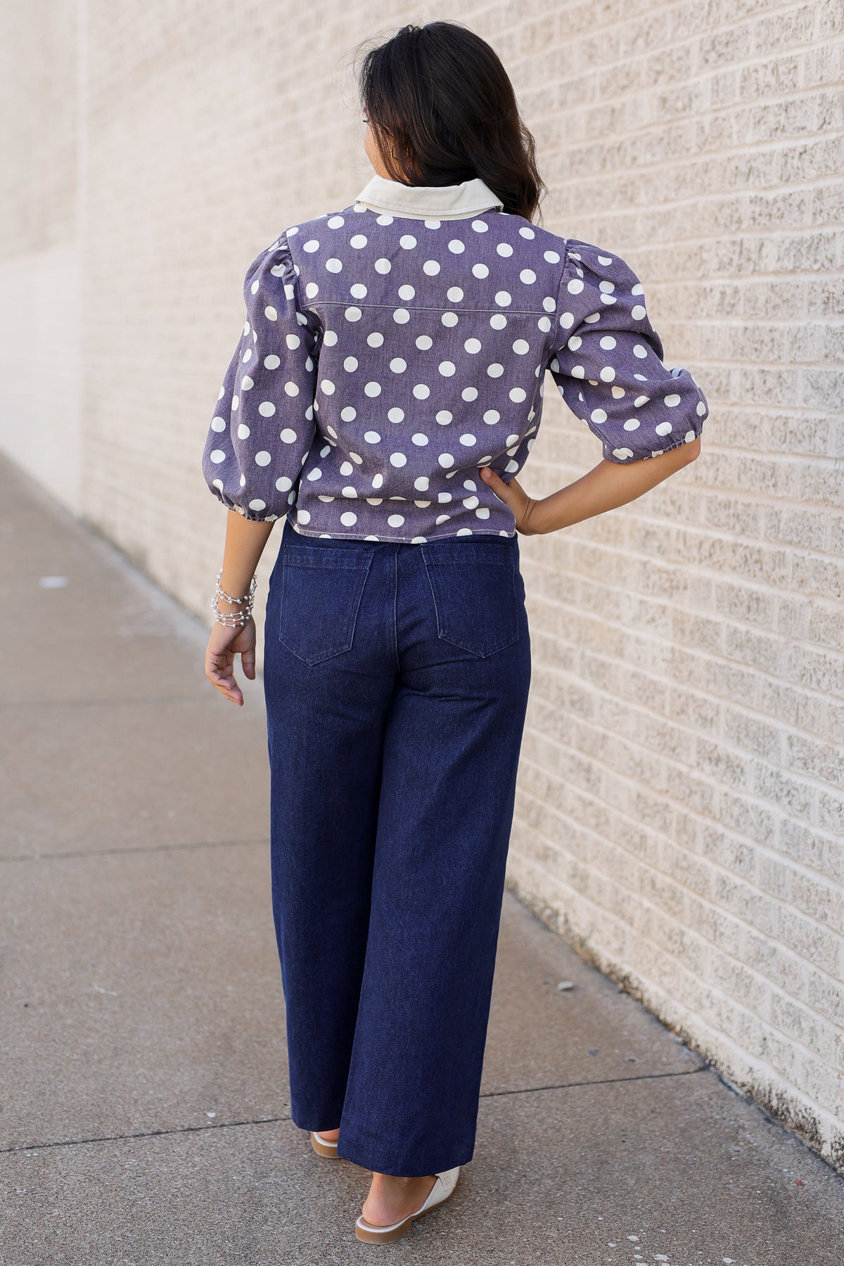 A woman viewed from behind wearing a lavender purple and white polka dot blouse with puffed sleeves and dark blue wide-leg jeans, standing on a sidewalk next to a light brick wall.