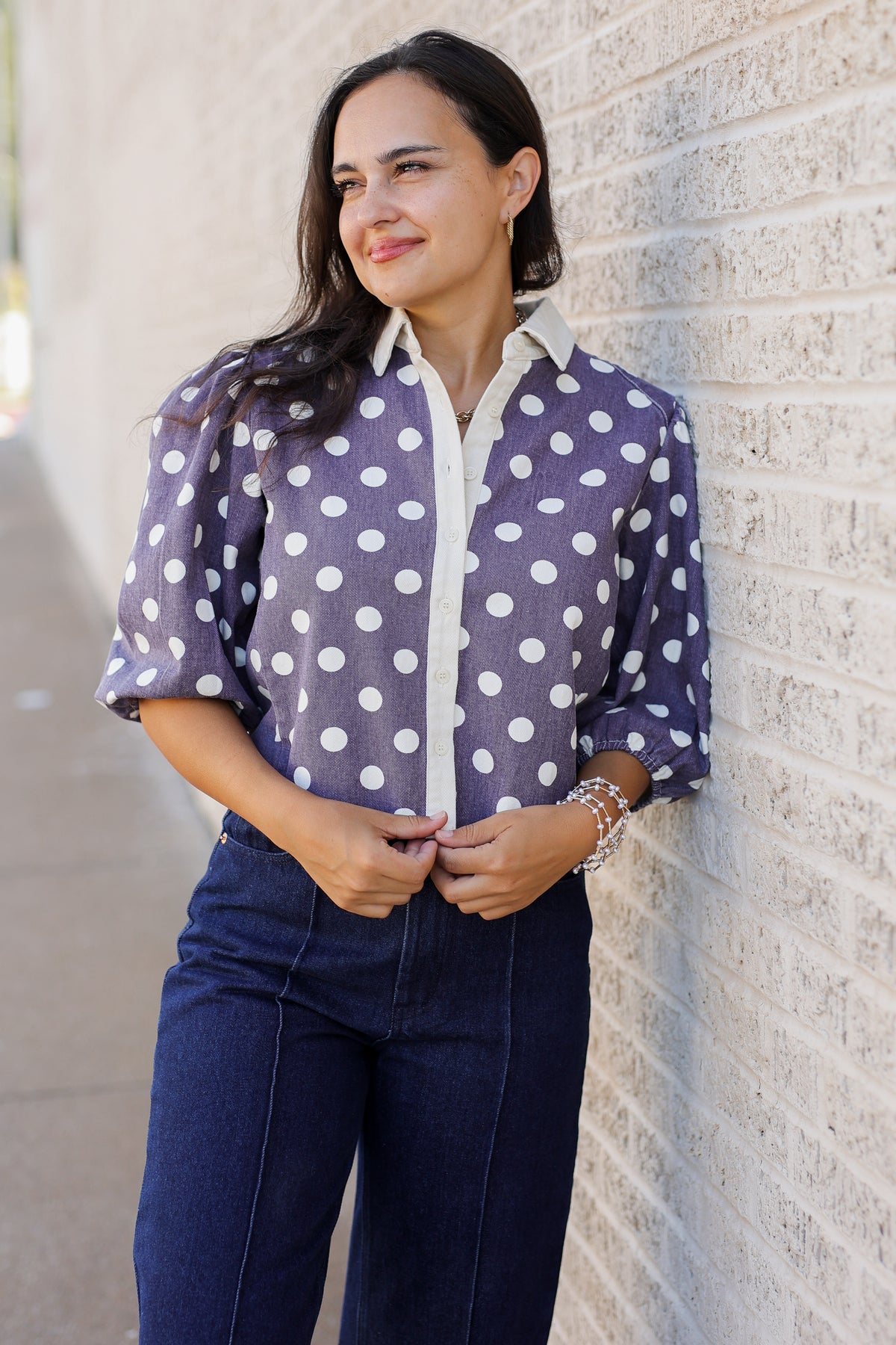 A woman wearing a purple and white polka dot blouse with dark blue jeans, standing against a light-colored brick wall.