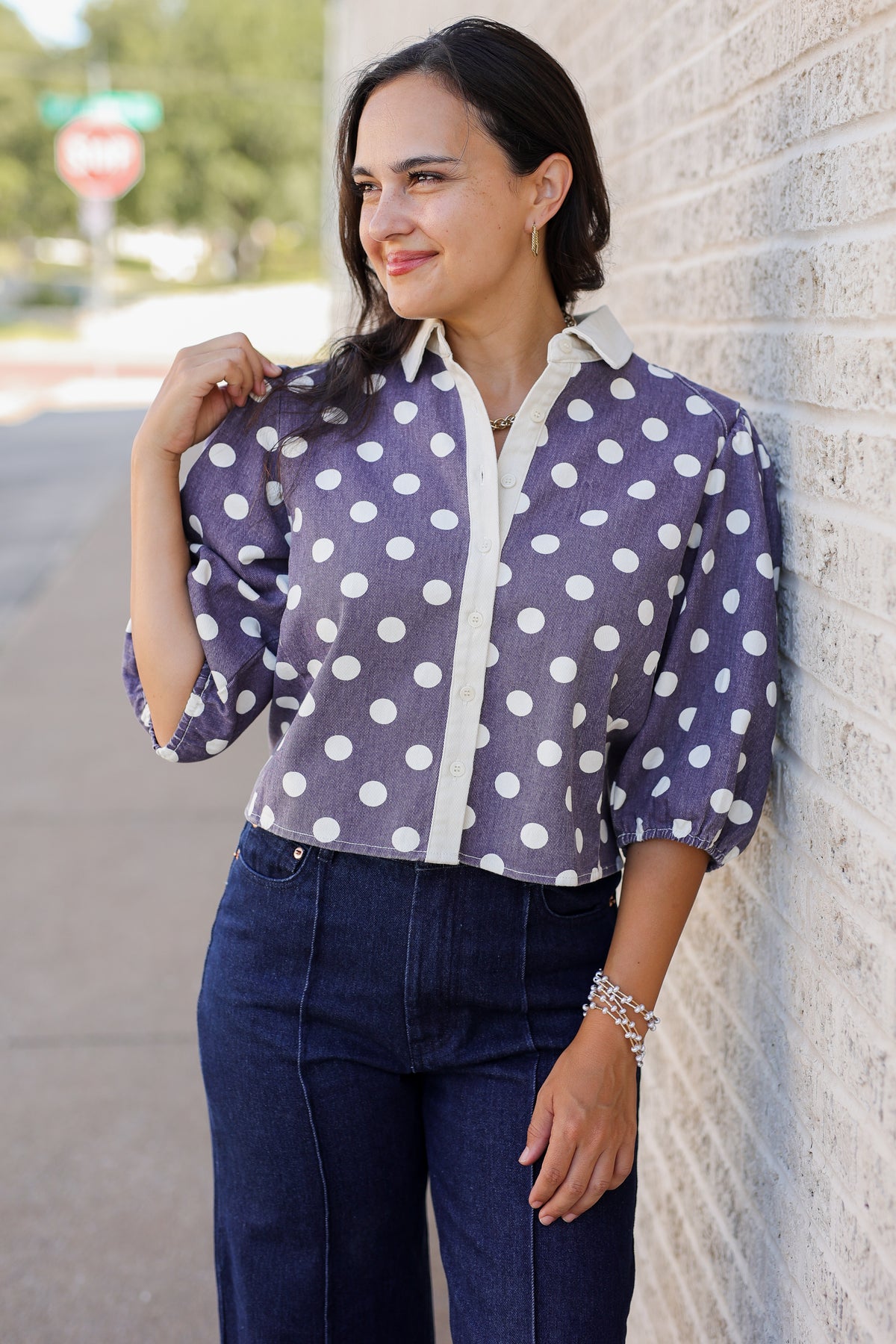 A woman wearing a purple and white polka dot blouse with three-quarter sleeves and dark blue jeans, standing outdoors against a light-colored brick wall.