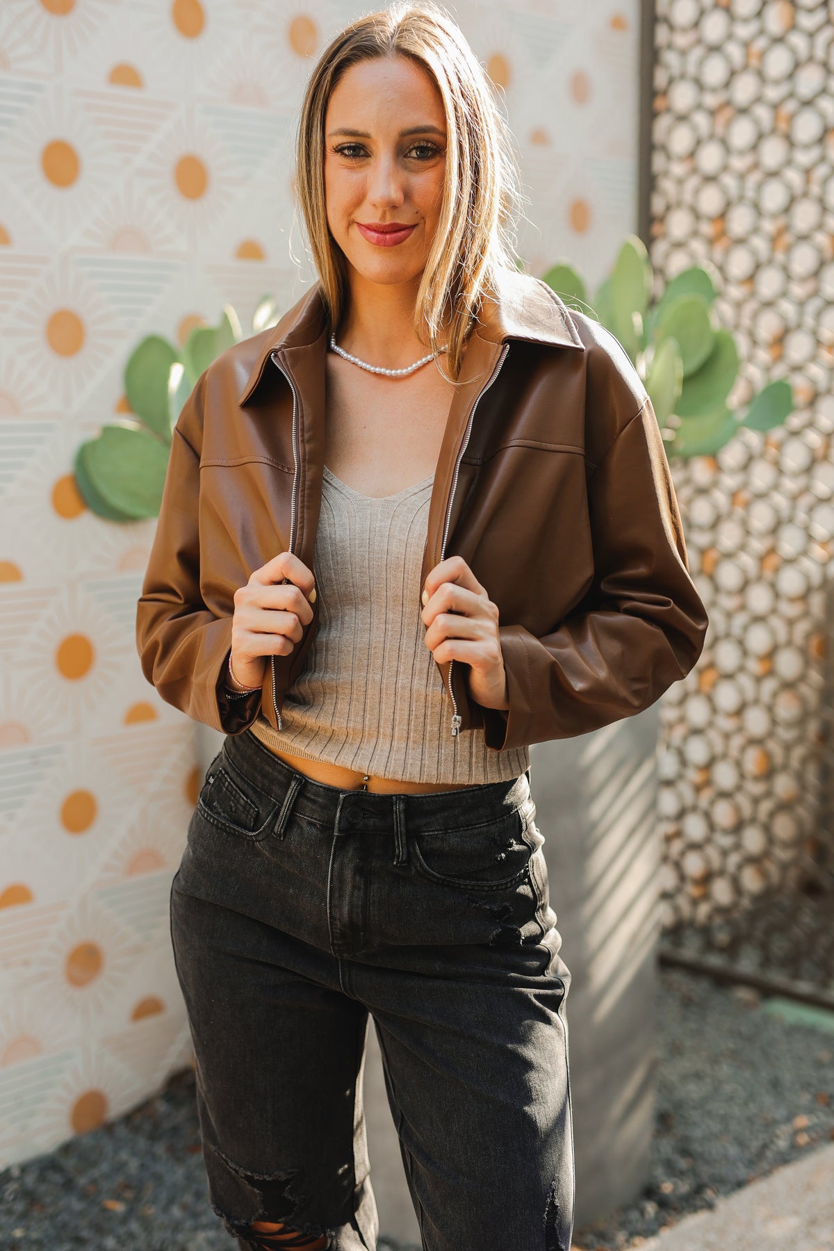 Woman wearing a brown jacket over a beige top and black pants, standing against a decorative wall.