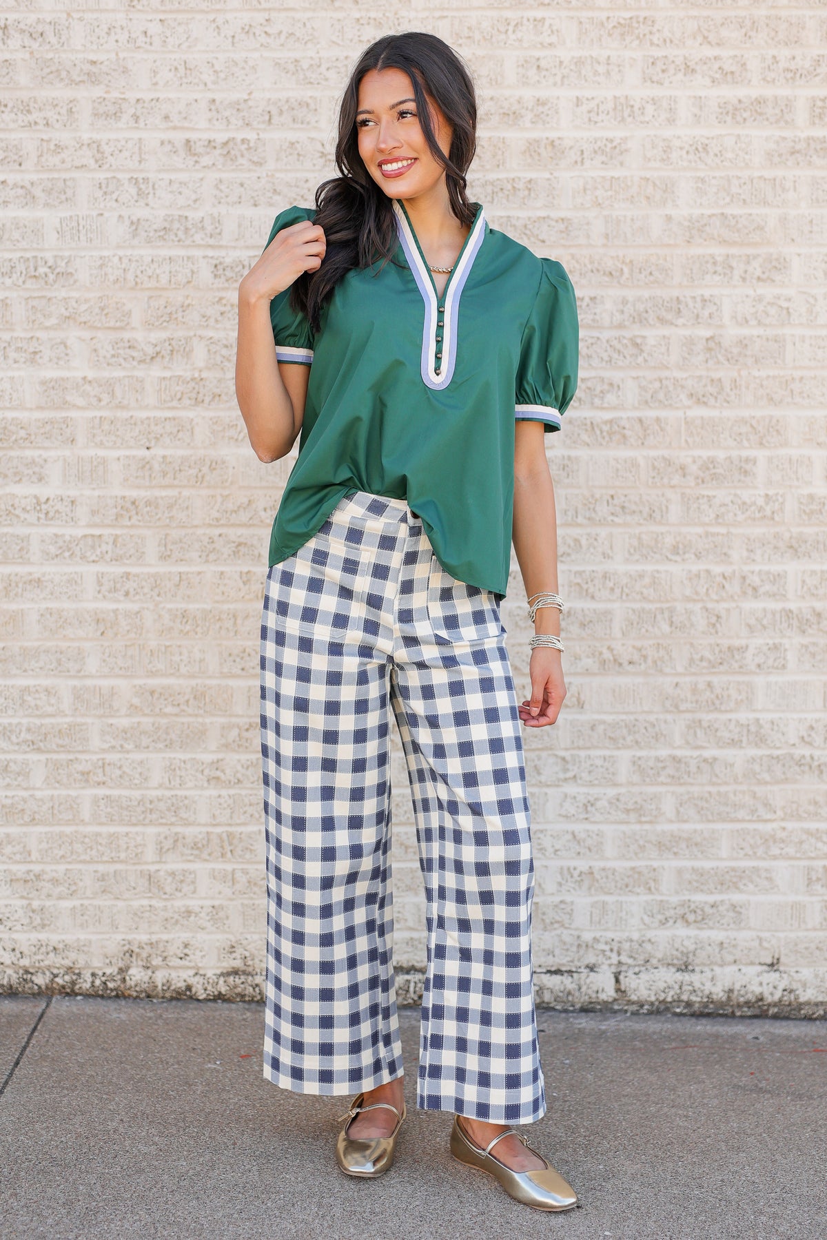 A woman modeling a green short-sleeve blouse with white trim and blue-and-white gingham wide-leg pants, standing against a light brick wall.