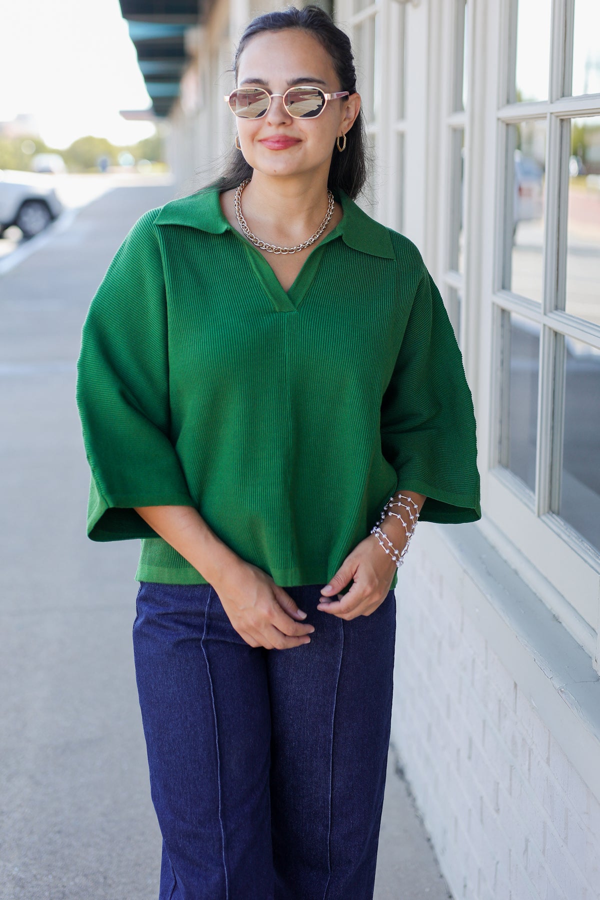 A woman wearing a vibrant green short-sleeve collared top and dark blue jeans, standing outdoors next to a white brick wall with large windows.