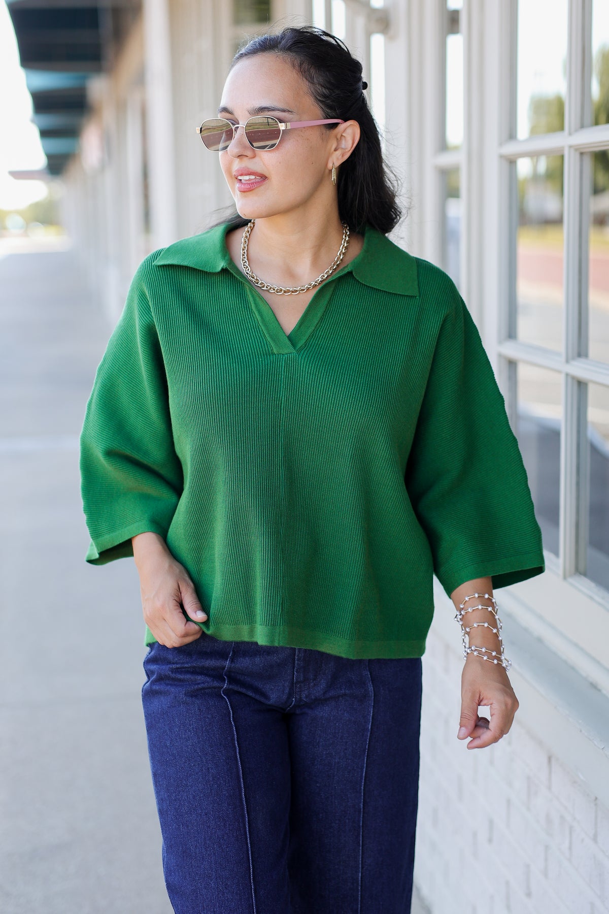 A woman wearing a vibrant green knit polo sweater and dark blue jeans, standing outdoors next to a white brick wall with large windows.