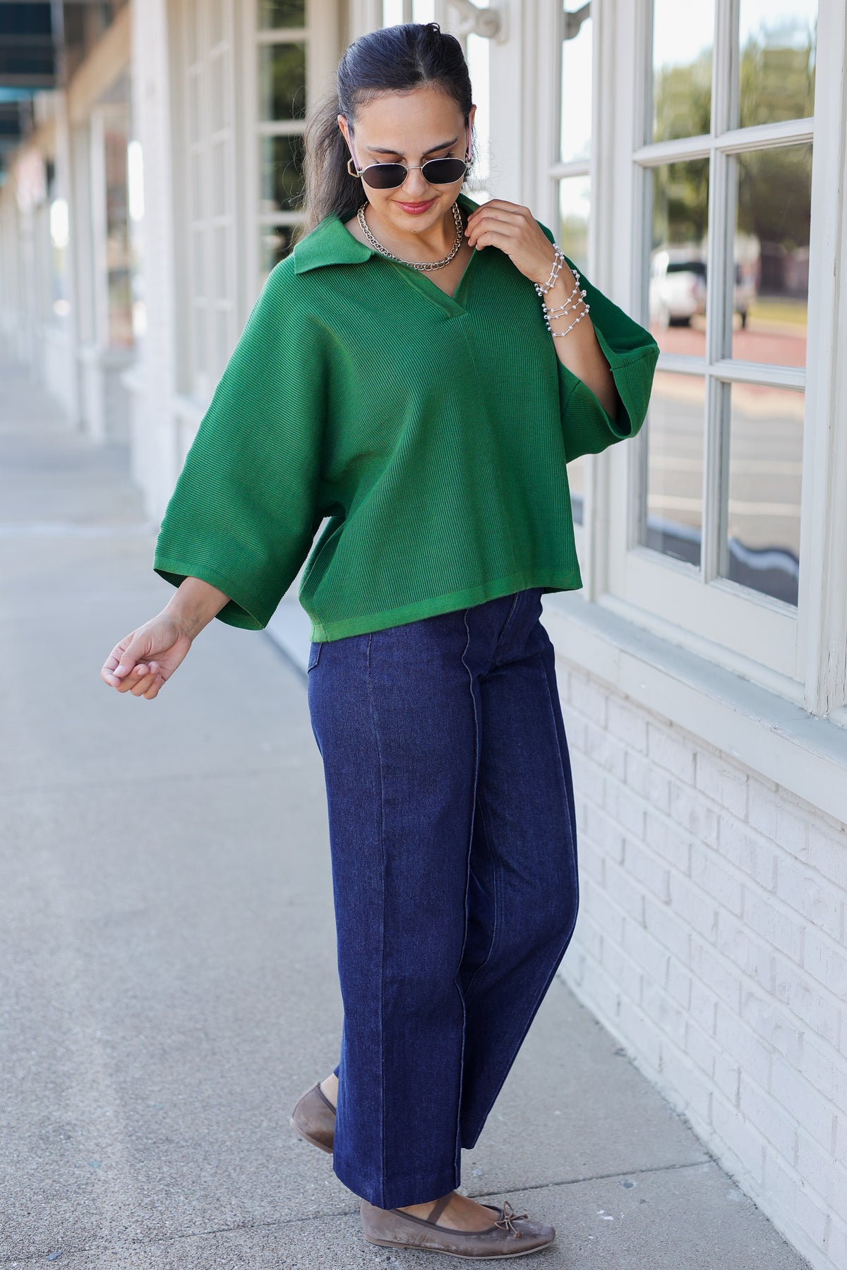 A woman wearing a vibrant green short-sleeve collared top and dark blue wide-leg pants, standing on a sidewalk next to a white brick building.