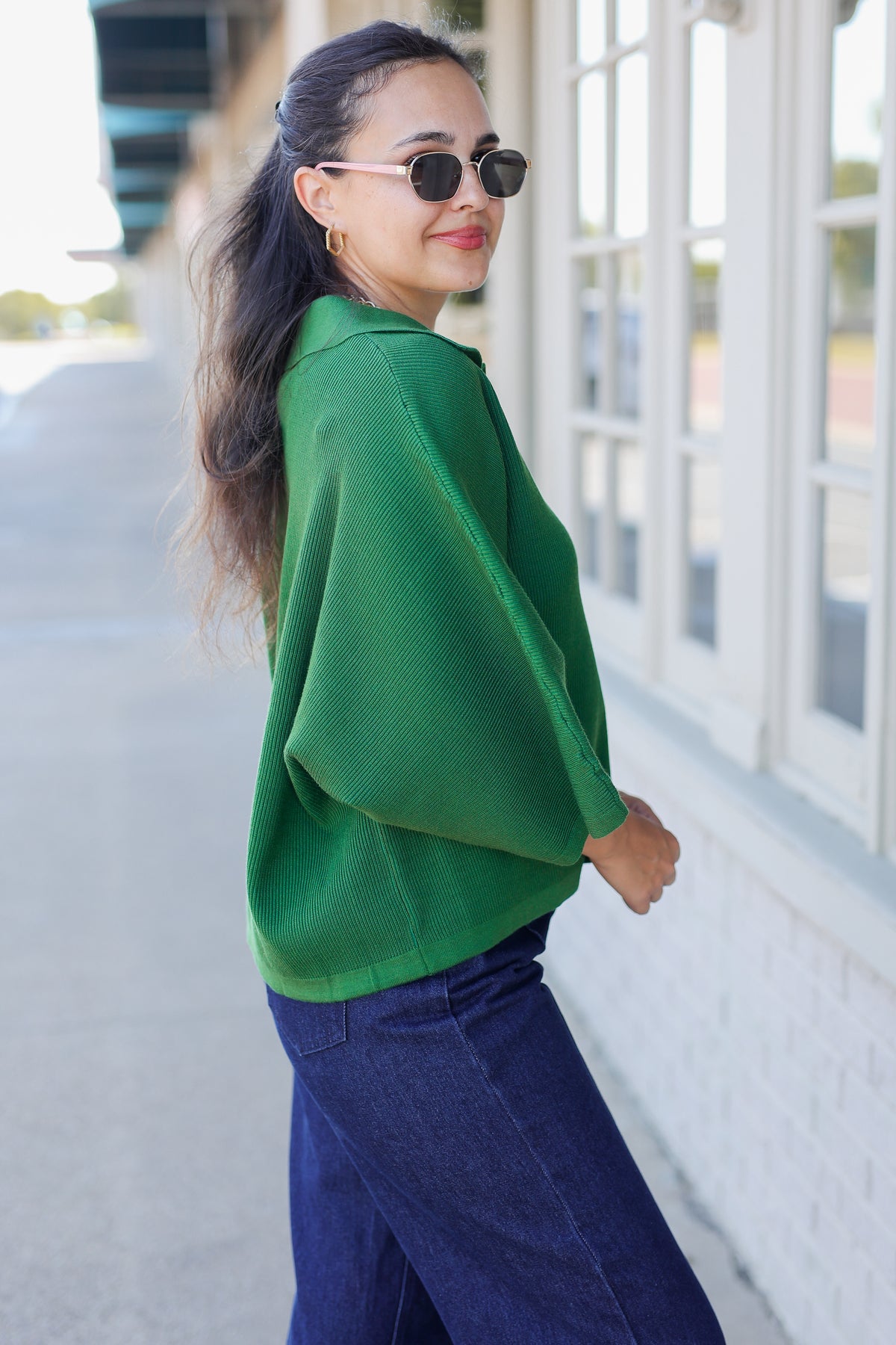 A woman wearing a vibrant green knit poncho-style top and dark blue jeans, standing outdoors next to a white building with large windows.