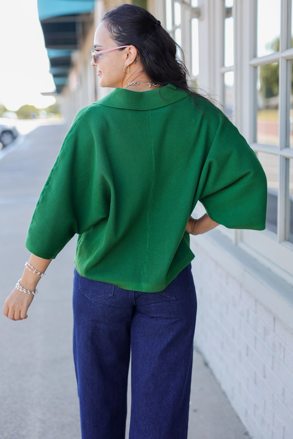 A woman wearing a bright green short-sleeve knit top and dark blue wide-leg pants, photographed from behind in an outdoor setting next to a building with large windows.