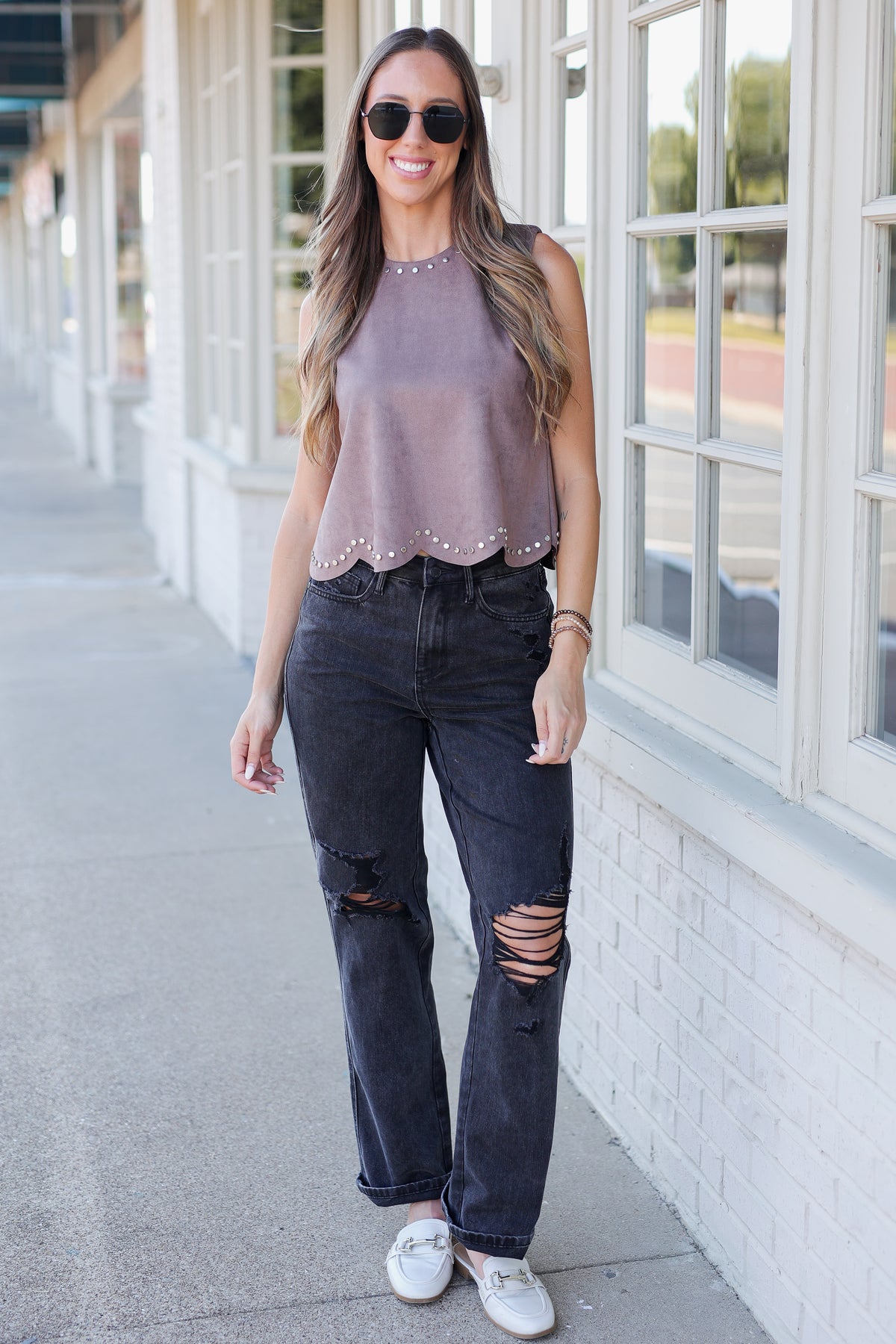 A woman wearing a sleeveless dusty mauve top with scalloped studded trim and dark jeans, standing outdoors in front of a building with large white-framed windows.