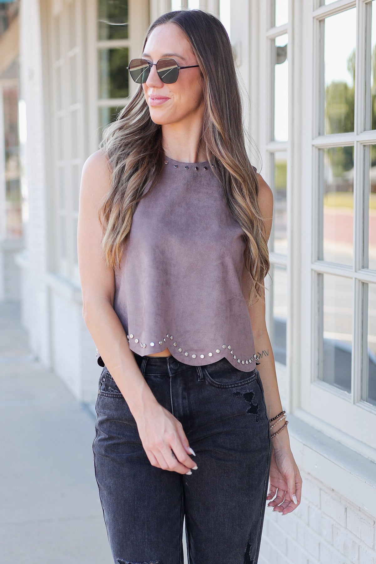 A woman wearing a sleeveless dusty mauve top with scalloped studded trim and dark jeans, standing outdoors in front of a building with large white-framed windows.