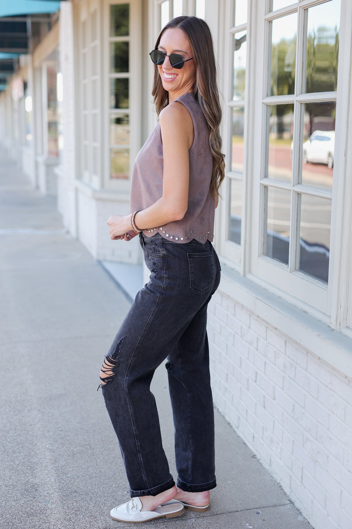 A woman wearing a sleeveless mauve top, dark washed distressed jeans, and white slide sandals stands on a sidewalk next to a white brick building with large windows.