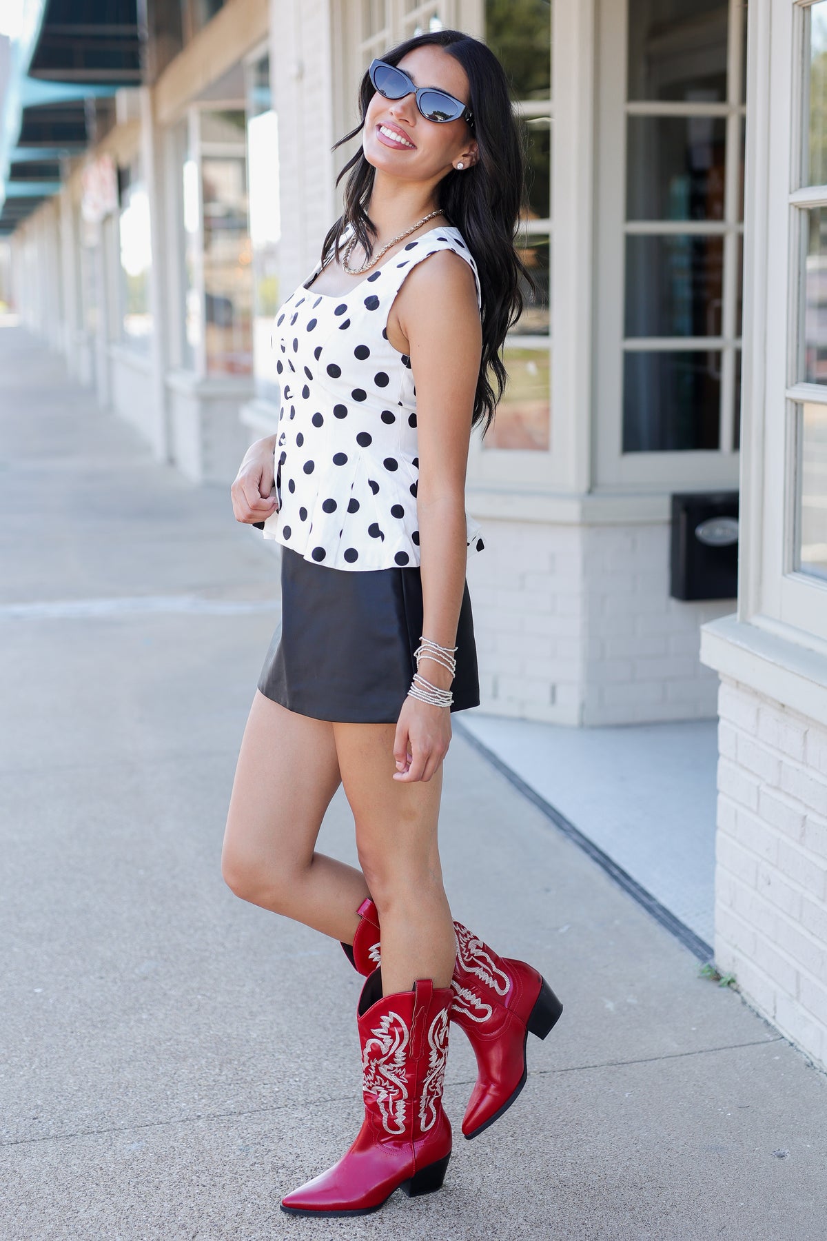 A woman posing on a sidewalk wearing a white sleeveless top with black polka dots, a black mini skirt, and red western cowboy boots with white embroidery.