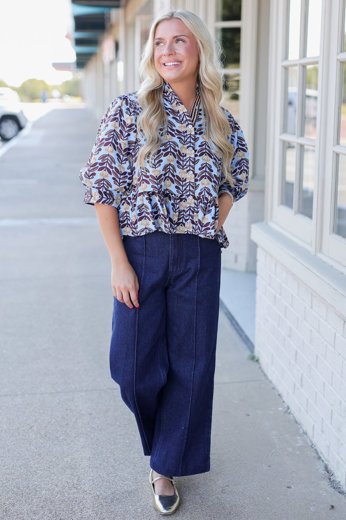 A woman stands on a sidewalk wearing a patterned blouse with three-quarter sleeves and dark wide-leg pants. The blouse has a multicolor abstract print with navy, burgundy, cream, and gold tones.