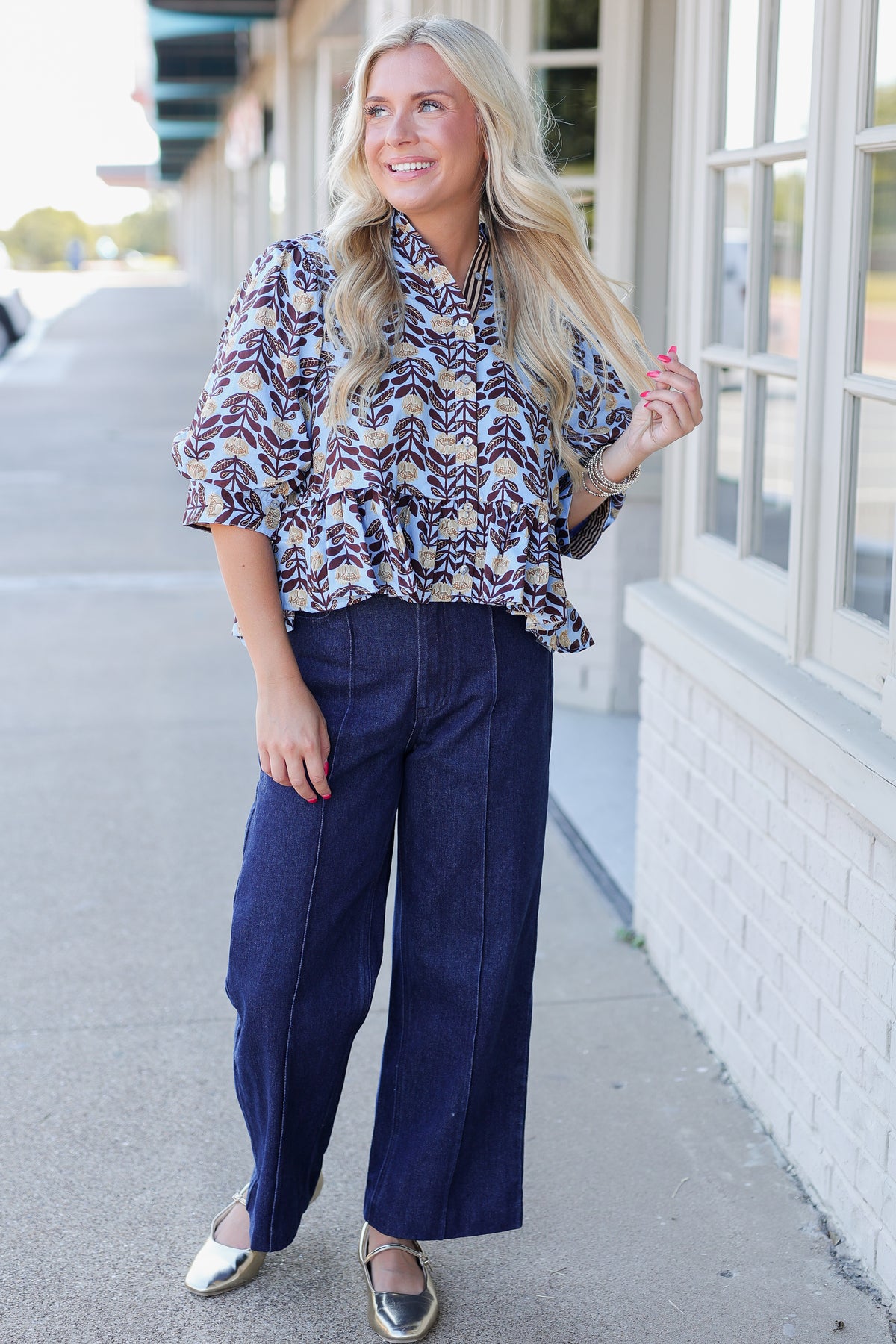 A woman stands on a sidewalk wearing a patterned button-up blouse with navy wide-leg pants and metallic flats, posing in front of a white brick building.