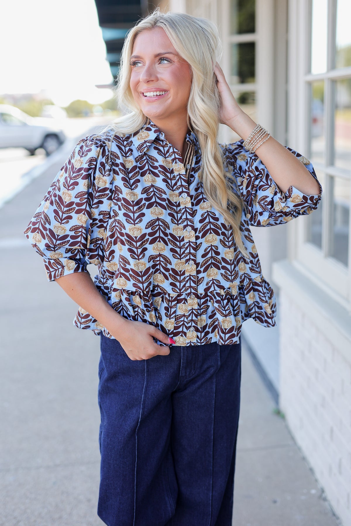 A woman with blonde hair stands on a sidewalk wearing a patterned button-up blouse with brown, beige, and light blue leaf-like motifs, paired with dark navy pants.