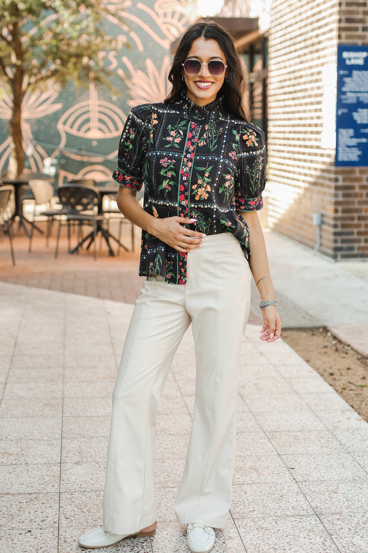 Woman wearing a black floral blouse and white pants standing outdoors.