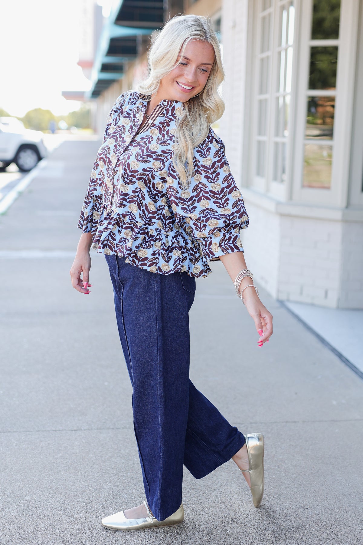 A woman walking outdoors on a sidewalk wearing a loose patterned blouse with purple, white, and beige tones, navy wide-leg pants, and metallic gold flats.