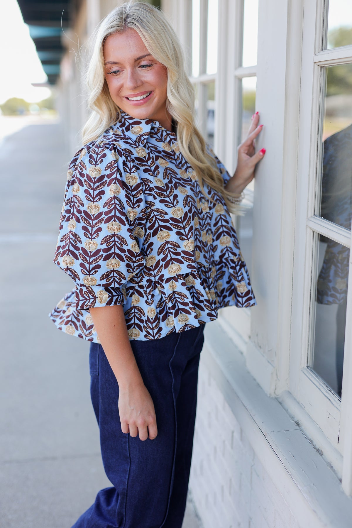 A woman with blonde hair stands outdoors next to a white building window, wearing a light blue blouse with a brown and beige leaf pattern and dark navy wide-leg pants.