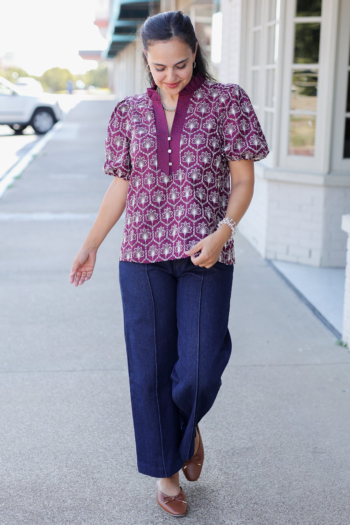 A woman walking outdoors on a sidewalk wearing a short-sleeve button-front blouse with a purple, pink, and white geometric pattern, paired with dark wide-leg pants and brown loafers.