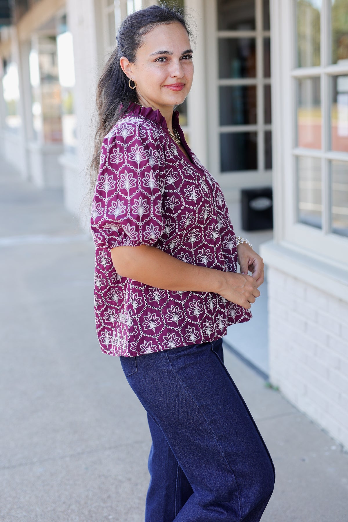A woman wearing a short-sleeve purple and white patterned blouse with dark blue jeans, standing outdoors in front of a storefront.