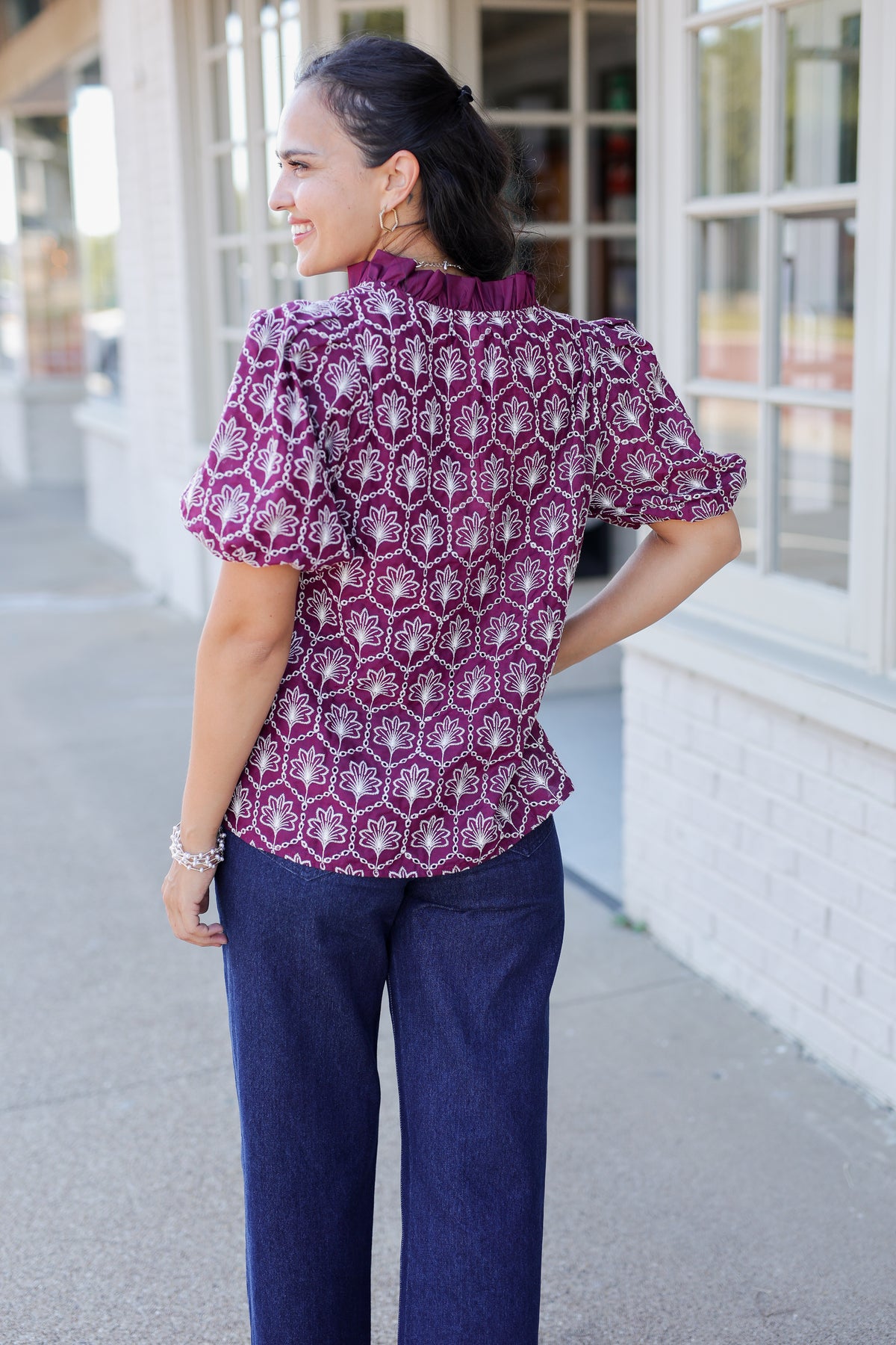 A woman photographed outdoors on a sidewalk in front of a building, wearing a short-sleeve patterned blouse with a high ruffled collar and dark blue pants, shown from the back.