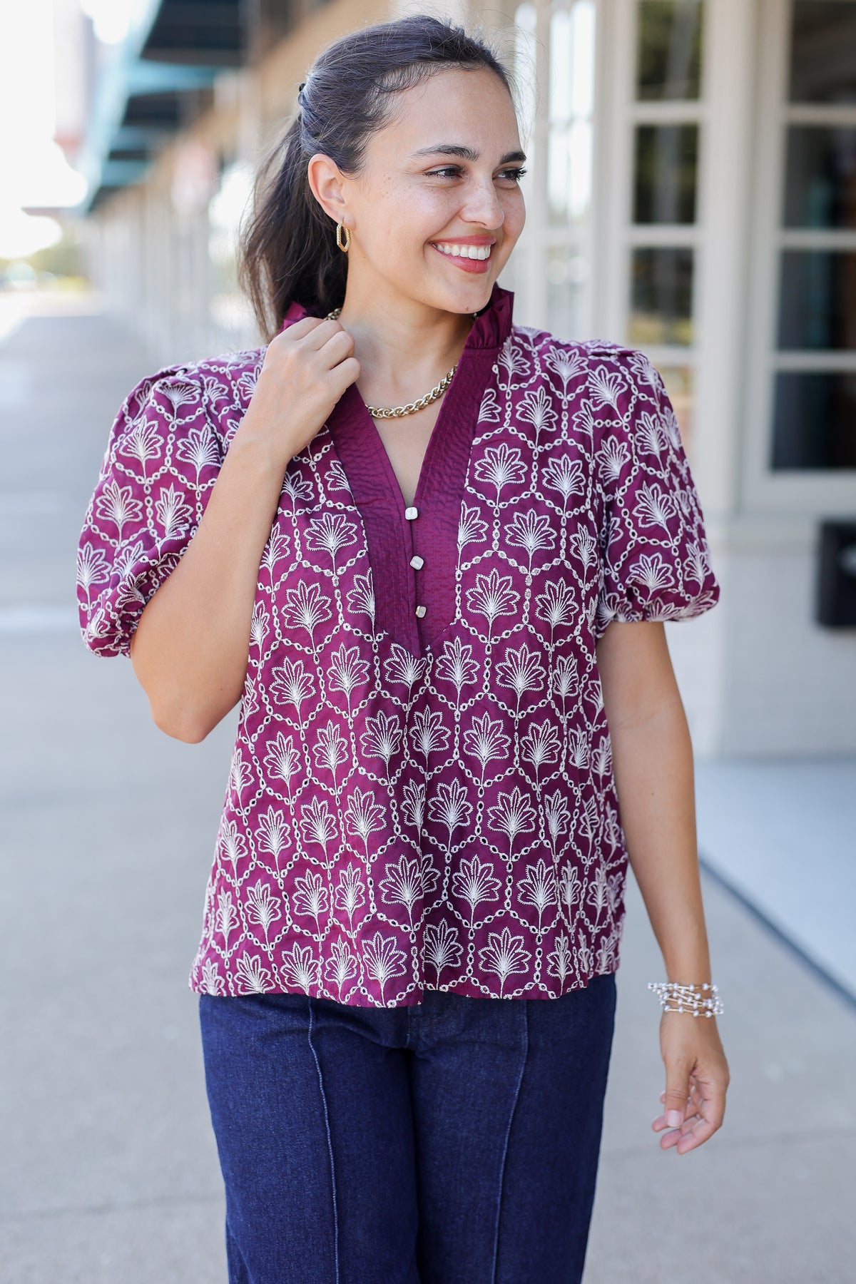 A woman wearing a short-sleeve button-front blouse with a purple and white geometric pattern, paired with dark blue jeans, standing outdoors on a sidewalk.