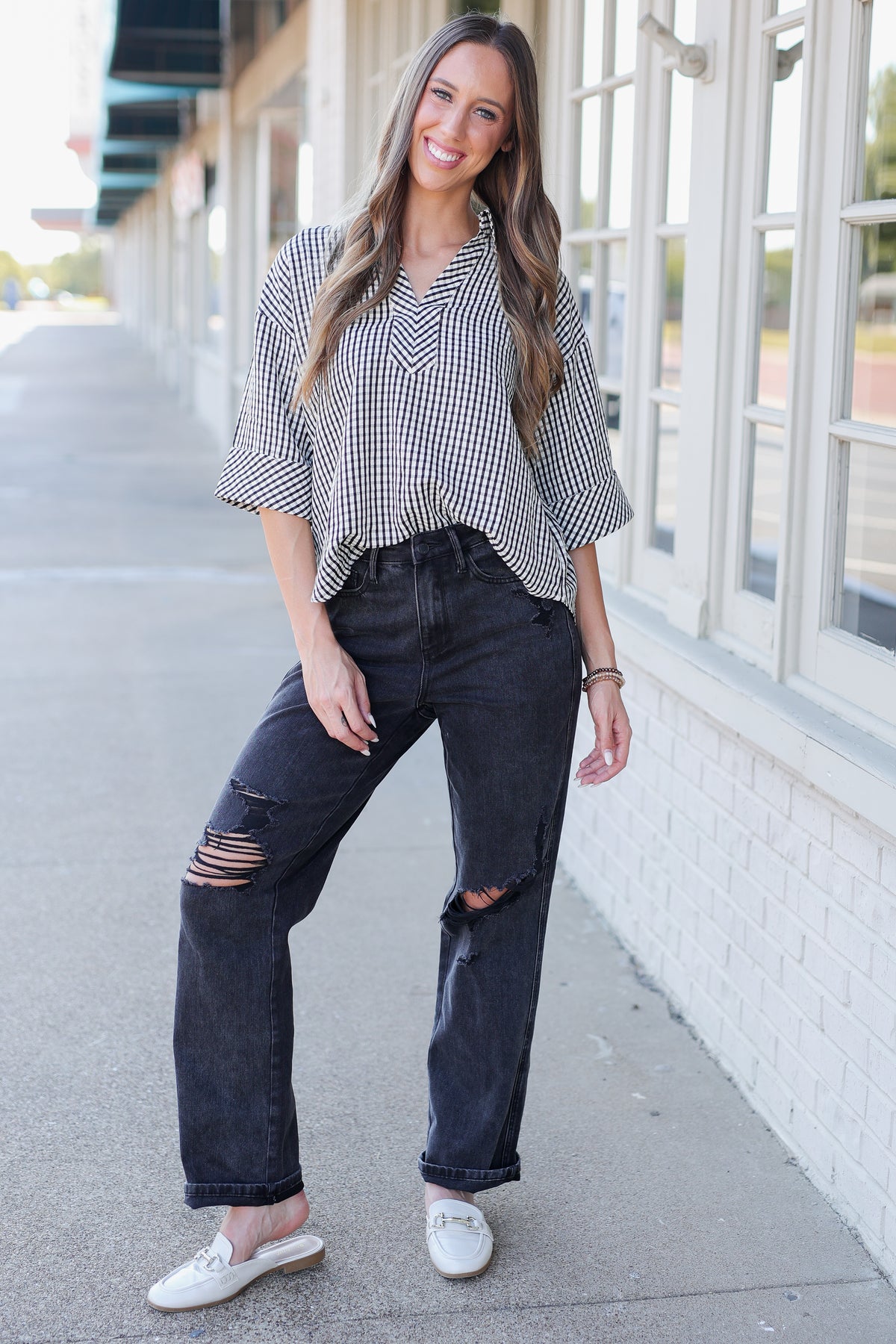 A woman stands on a sidewalk wearing a black and white gingham patterned blouse with three-quarter sleeves, dark distressed jeans, and white slip-on shoes.
