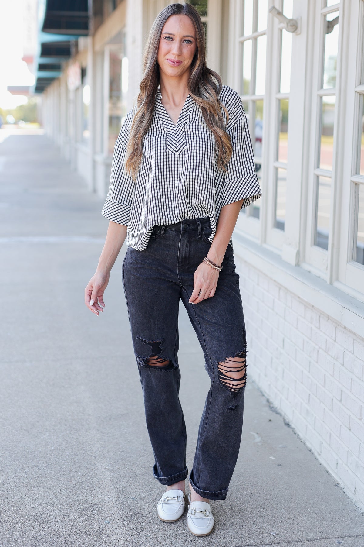 A woman stands on a sidewalk wearing a black and white gingham button-down shirt, dark distressed jeans, and white loafers, with storefront windows in the background.